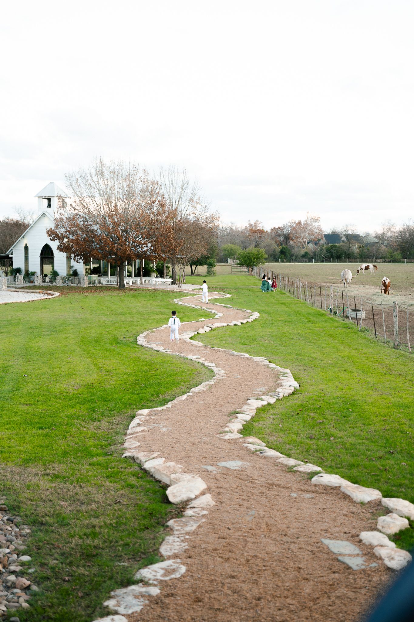 A winding stone-lined dirt path leads through a green lawn towards a white church building in the distance.