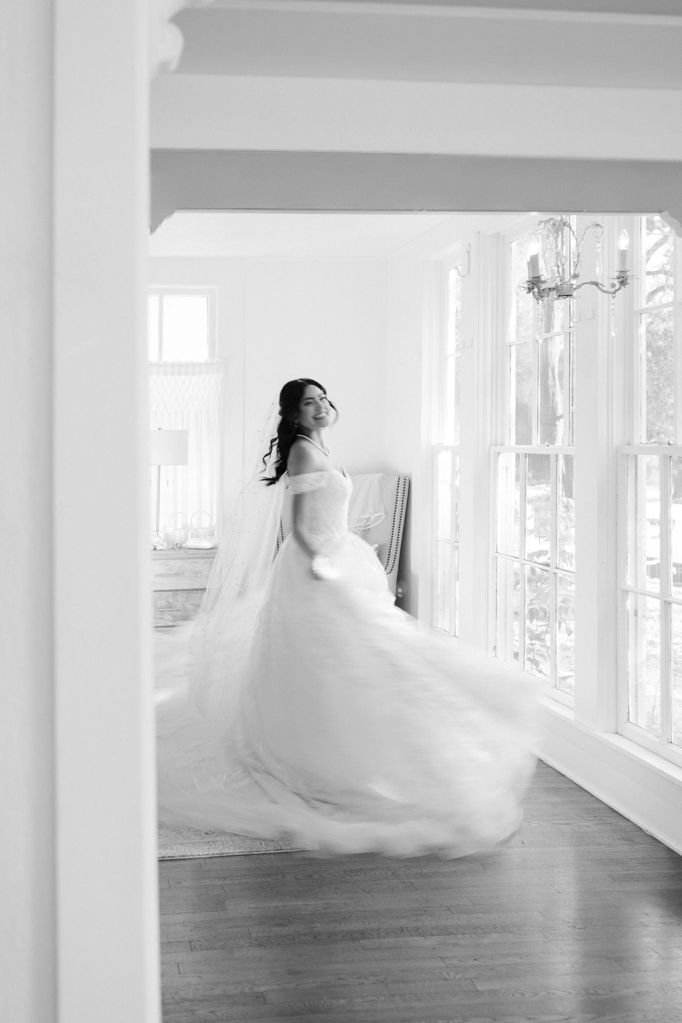 A bride in a flowing white wedding gown twirls gracefully in a bright sunlit room with hardwood floors and white walls.