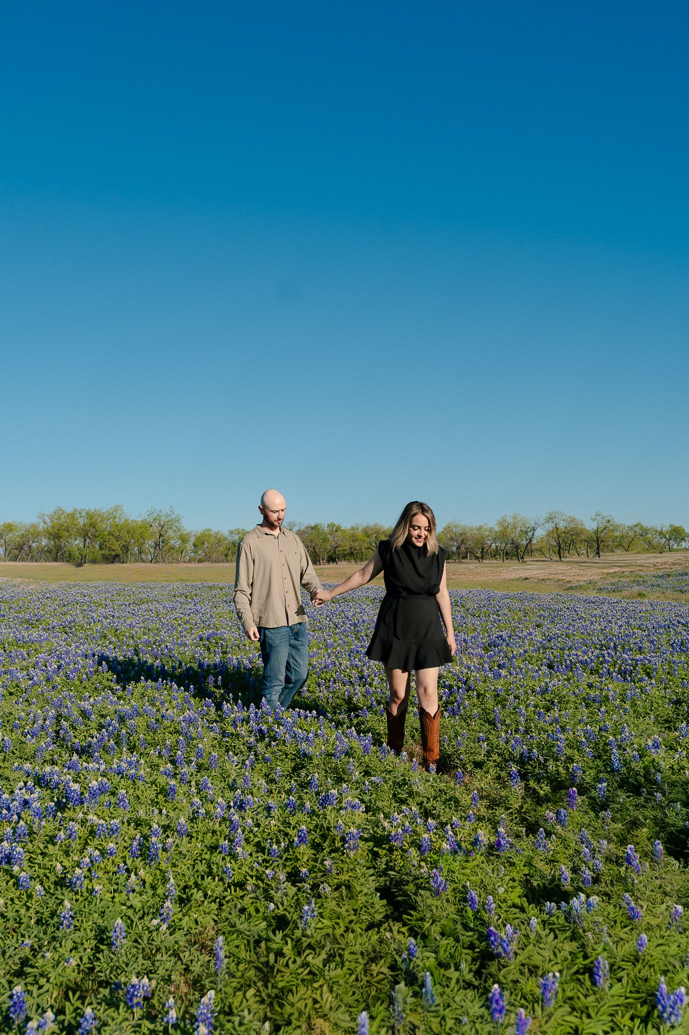 A couple walks through a vibrant field of blooming Texas bluebonnets under a bright blue spring sky.