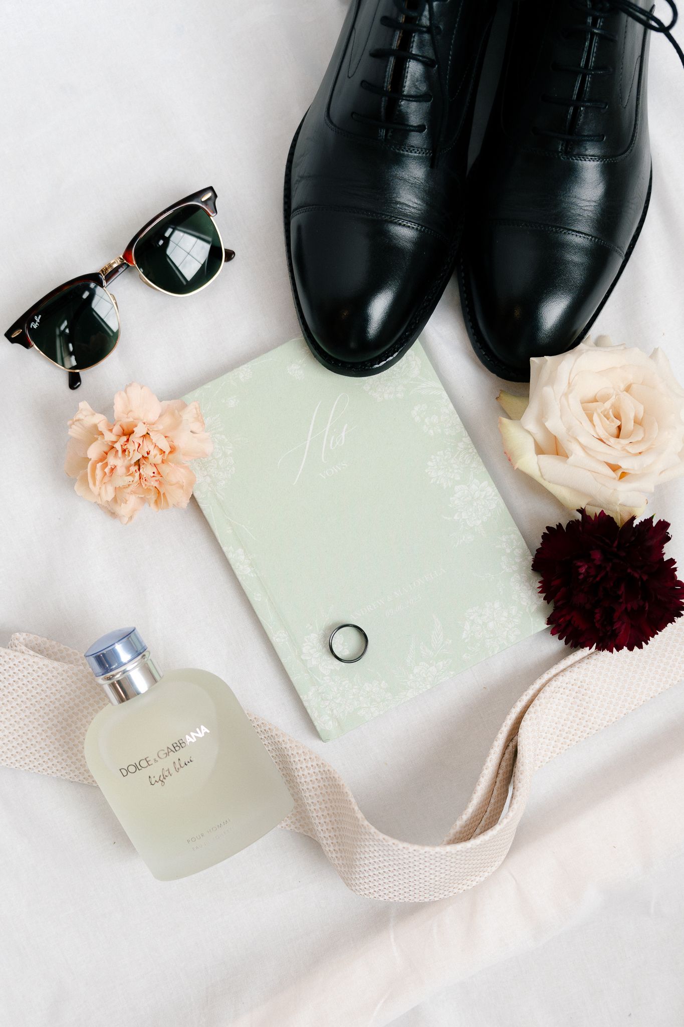 A flat lay of black leather dress shoes paired with delicate peach roses and burgundy flowers on a white surface.