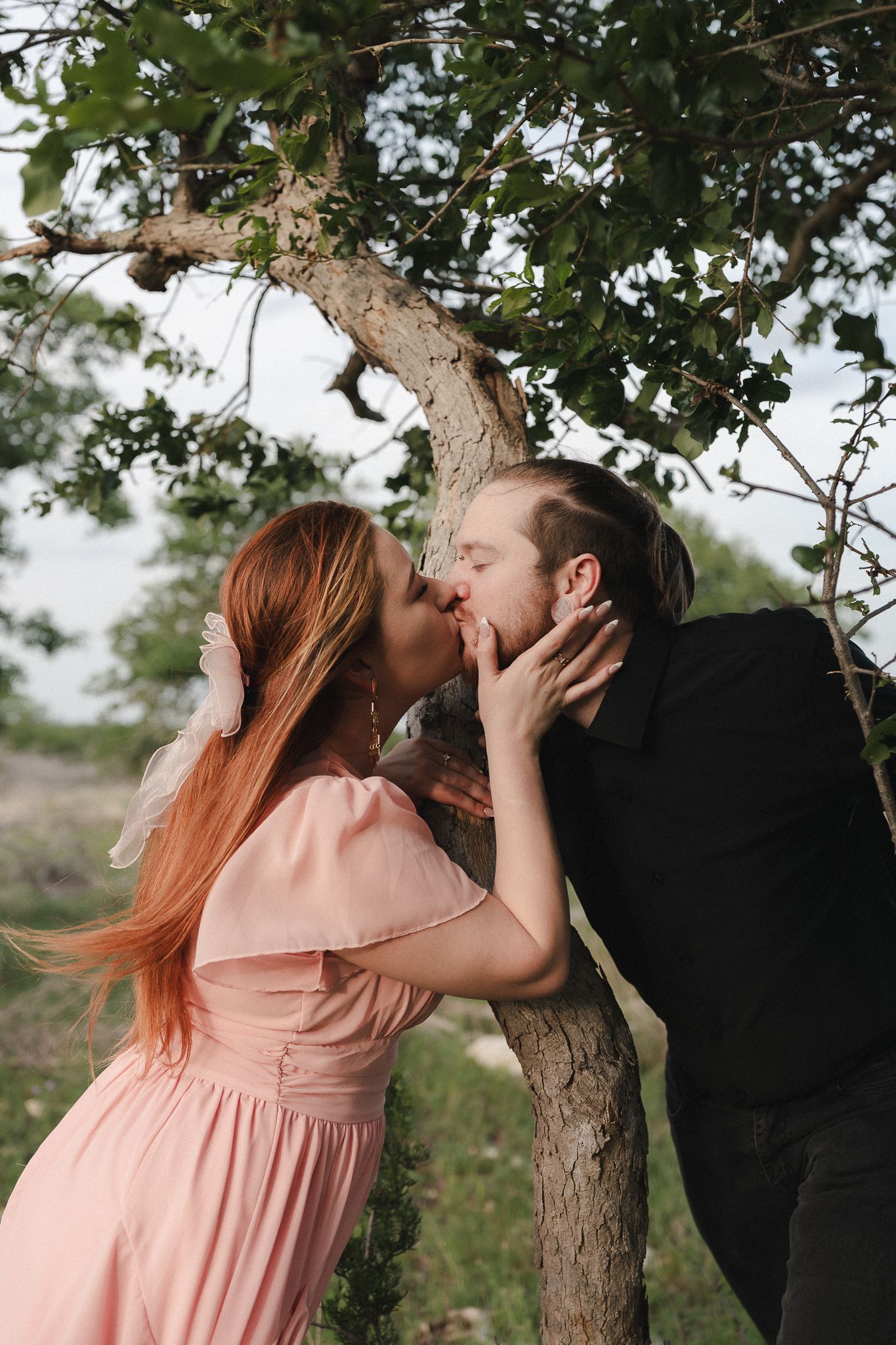 Romantic couple sharing a kiss under a tree, woman in pink dress and man in black suit outdoors.