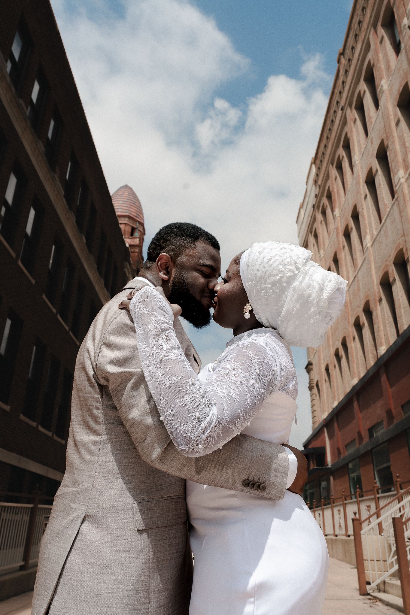 Black couple in wedding attire sharing a kiss outdoors between urban buildings on a sunny day.