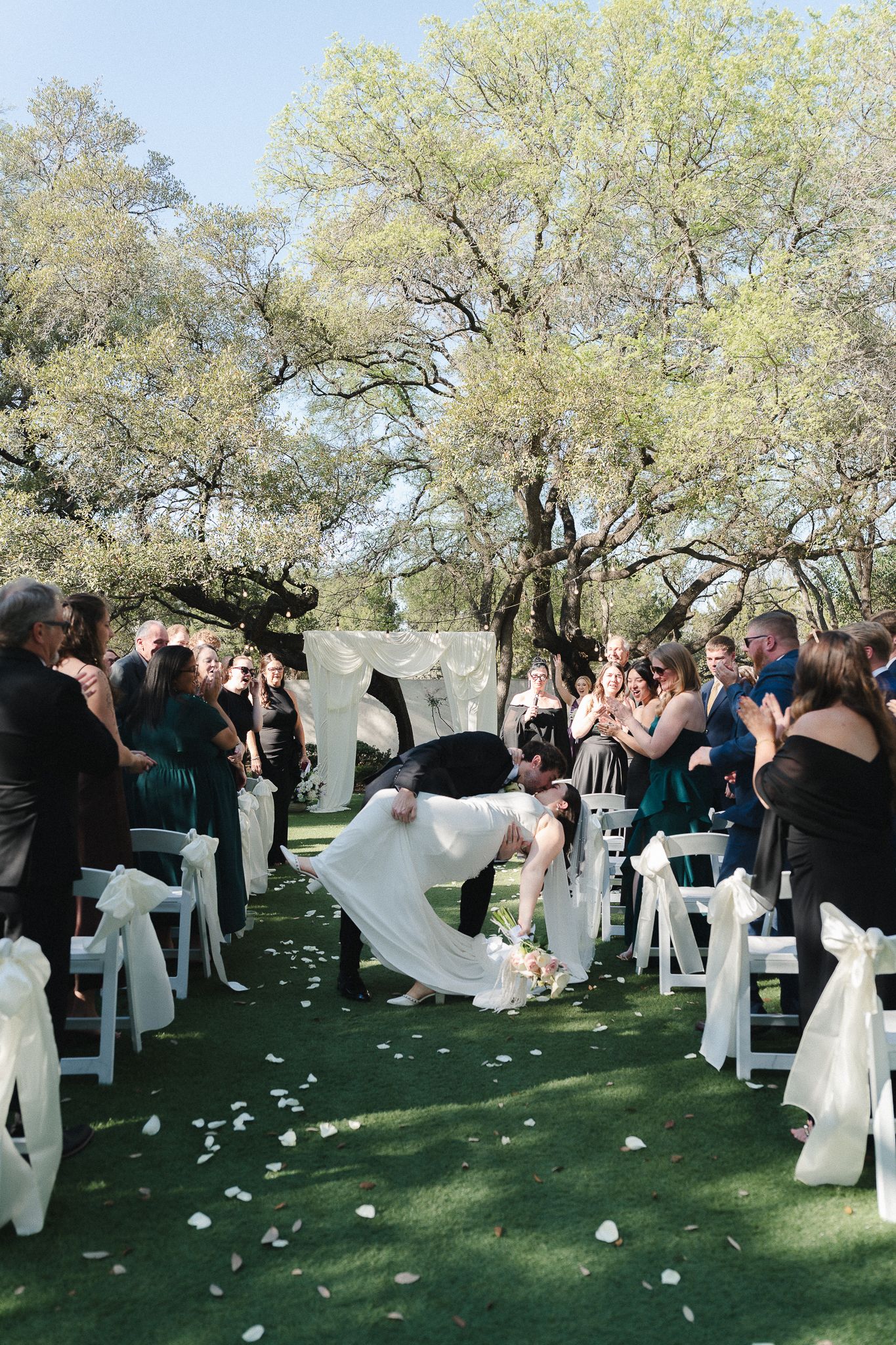 Groom dips bride for a romantic kiss at outdoor wedding ceremony with white chairs and rose petals on green lawn.