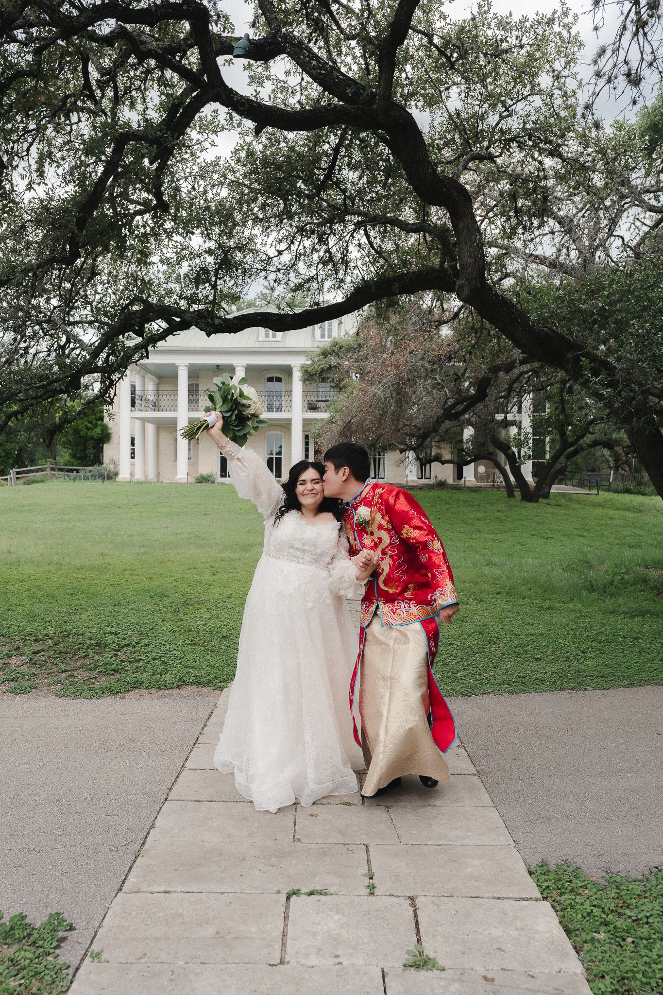 Bride in white gown and groom in red Chinese traditional attire share a kiss on a garden path outside a white mansion.