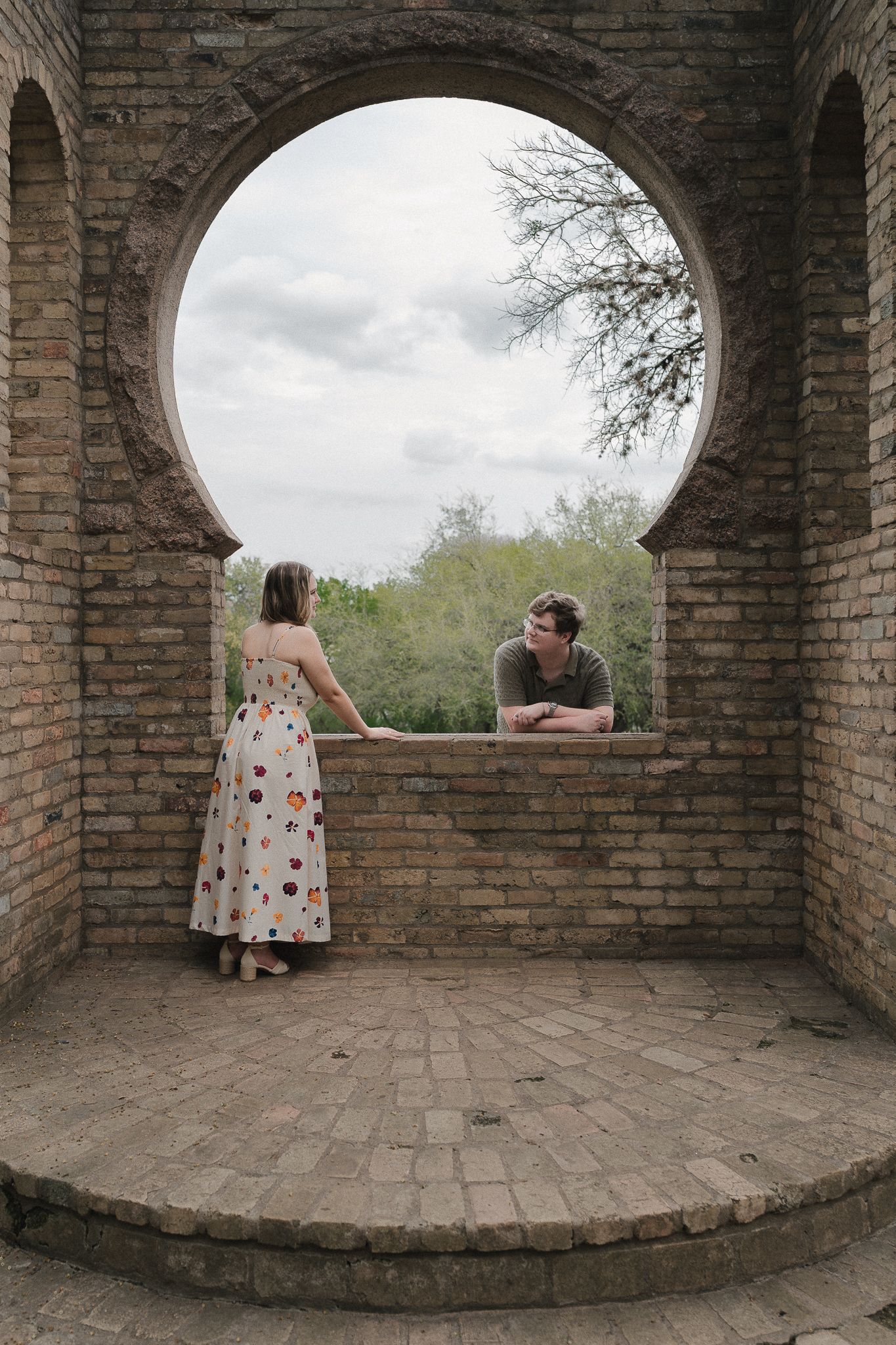 Couple sharing a romantic moment at a circular brick archway window, woman in floral dress holding hands with man leaning over wall.