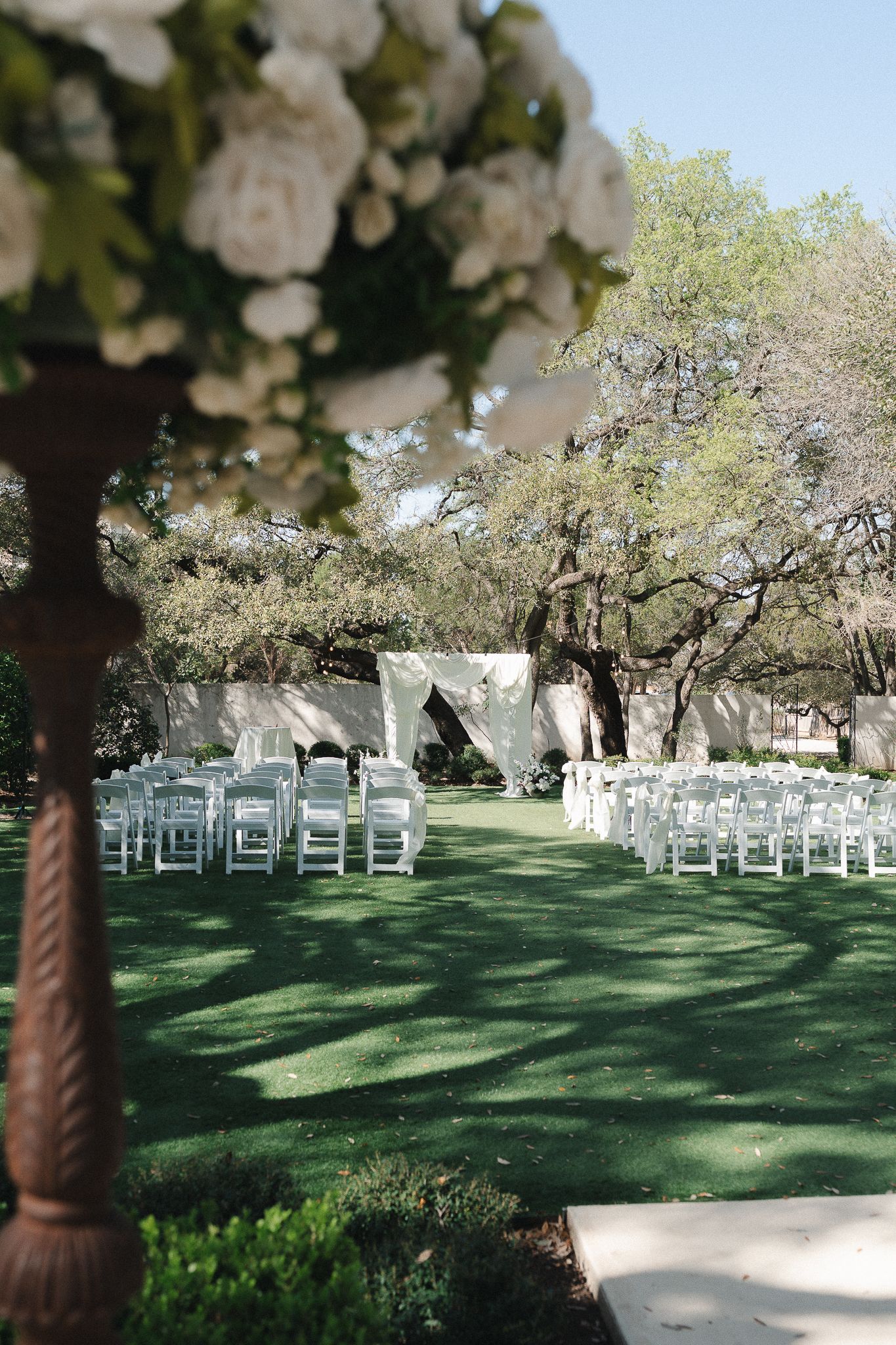 Outdoor wedding ceremony setup with white chairs on green lawn, floral arch backdrop, and large oak trees under clear blue sky.