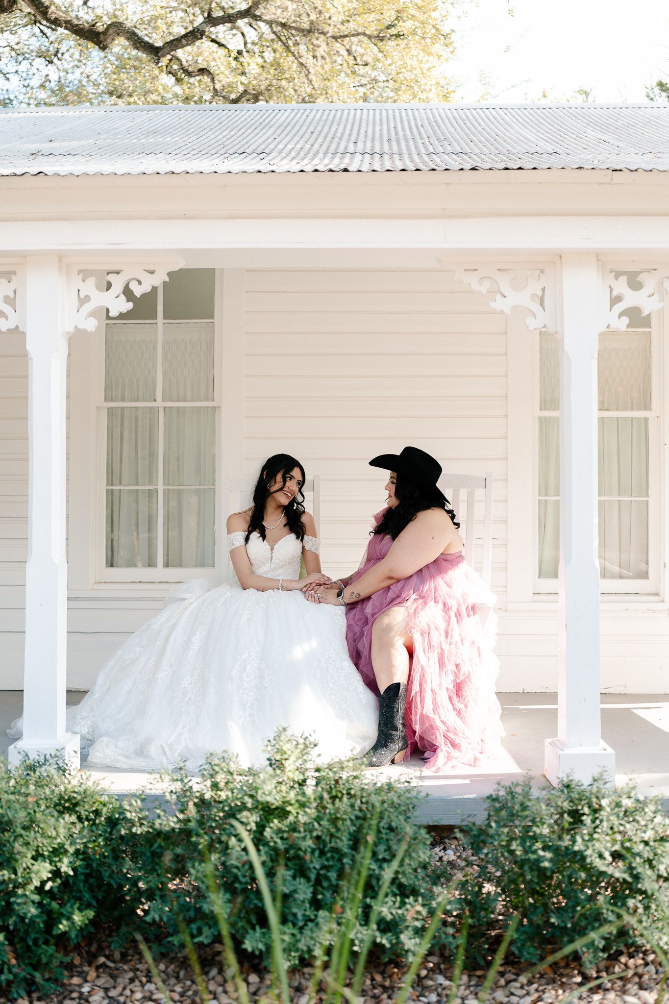 Two people share a sweet moment on a white farmhouse porch, one in a wedding dress and one in pink.