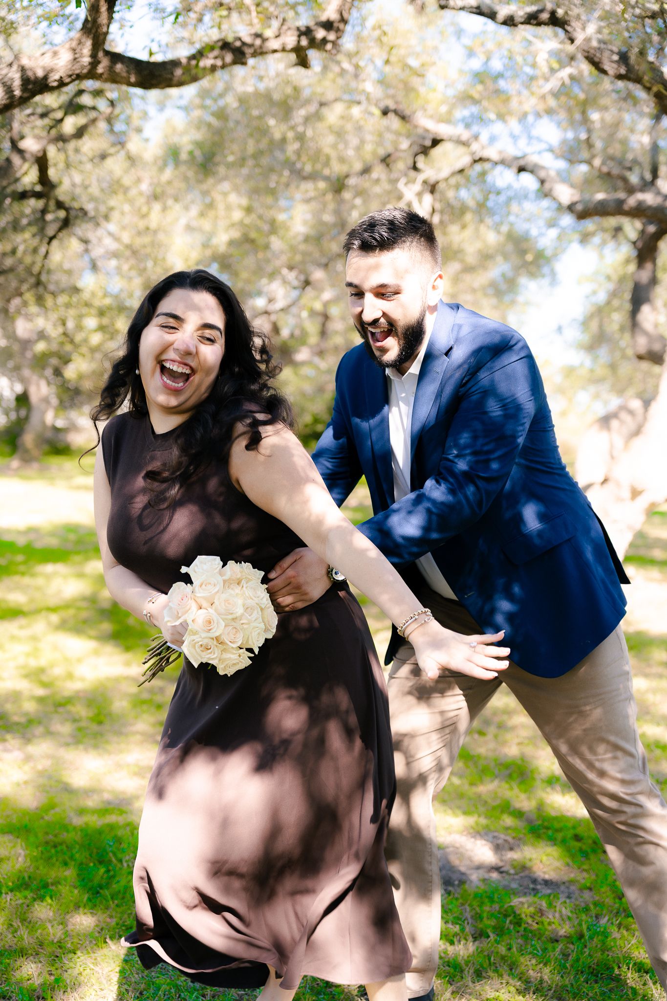 A joyful couple laughs together outdoors under a tree while holding a white bouquet.