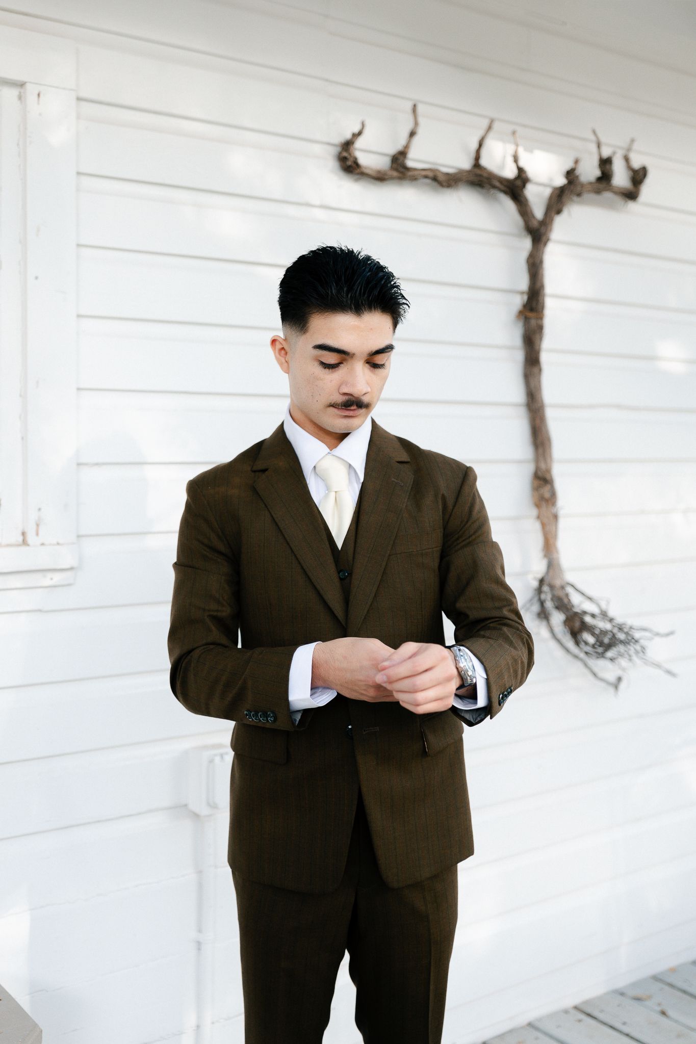 Person in a brown suit stands against a white wall in a classic vintage-style pose.