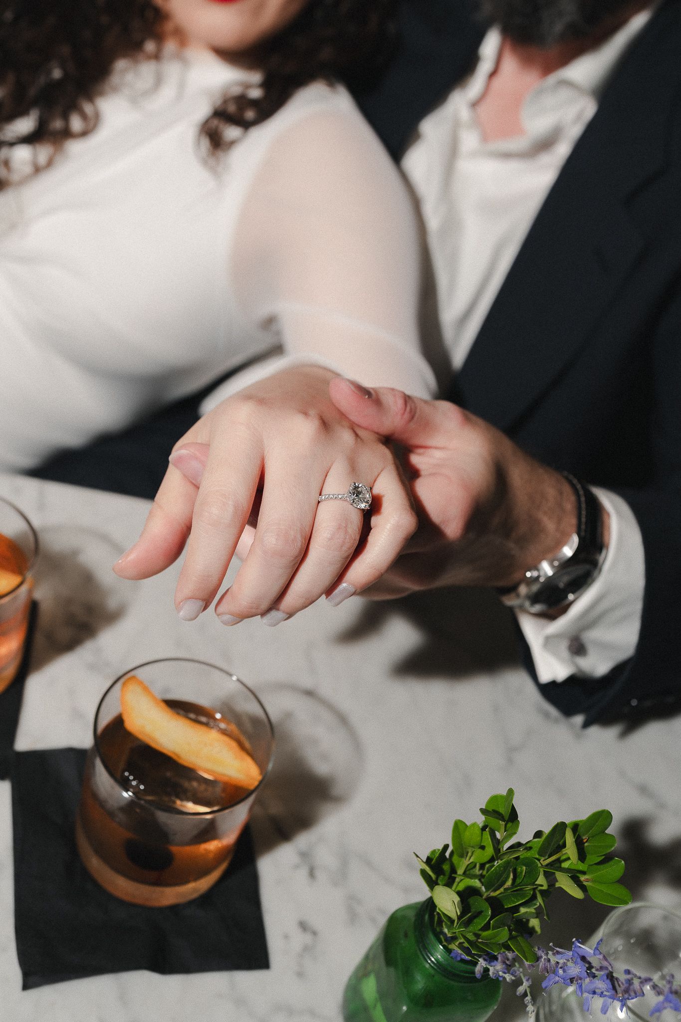 Couple at marble table showing off diamond engagement ring, with cocktails garnished with orange slices nearby.