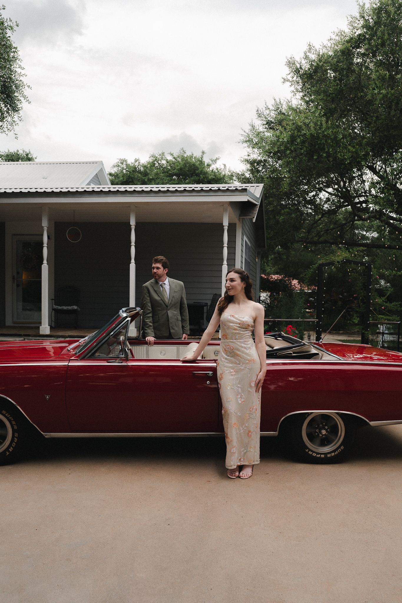 Couple in formal attire posing by a vintage red convertible in front of a charming house with a porch.