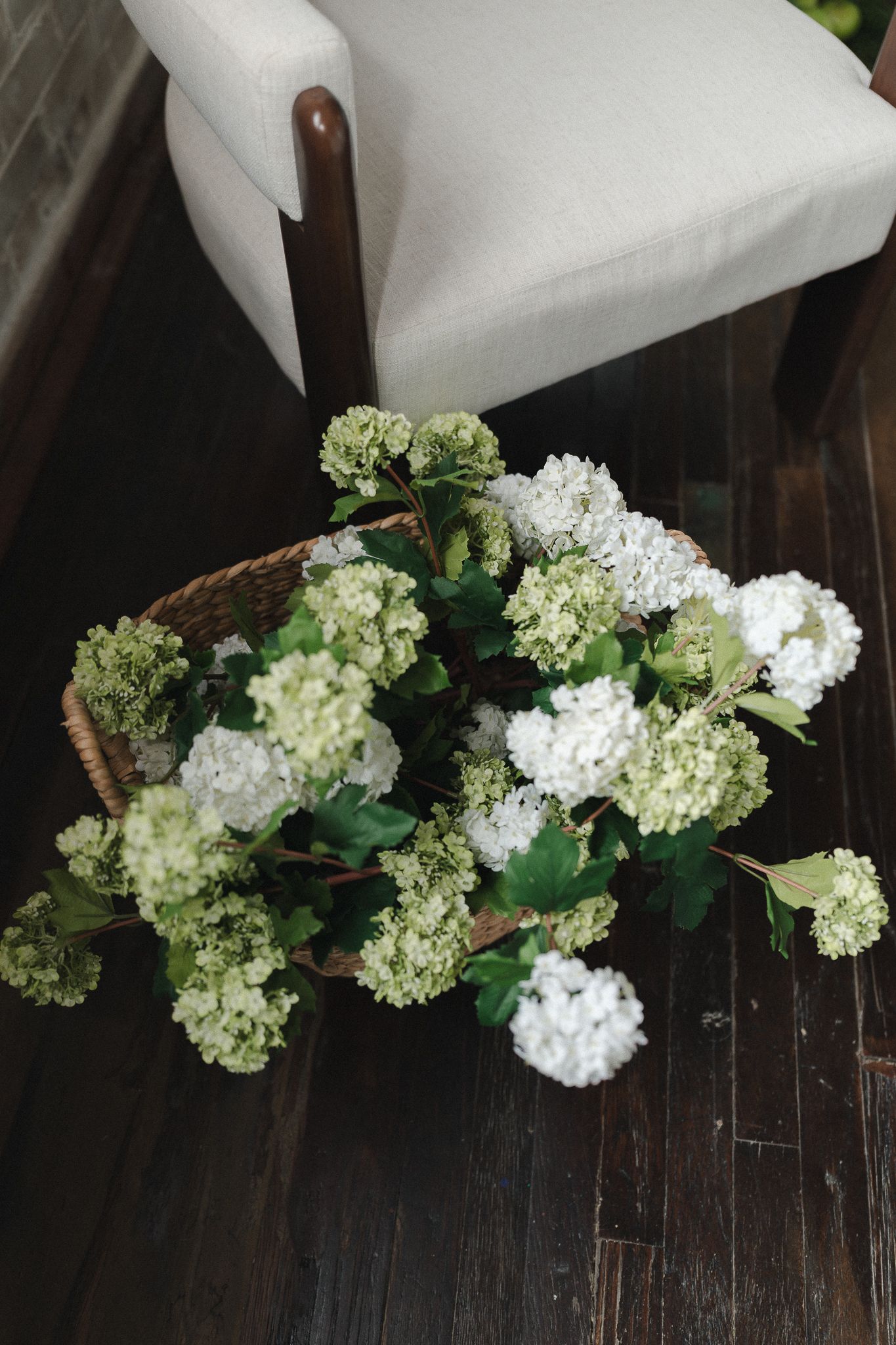 Wicker basket filled with white and green hydrangea blooms on dark hardwood floor beside a white upholstered chair.