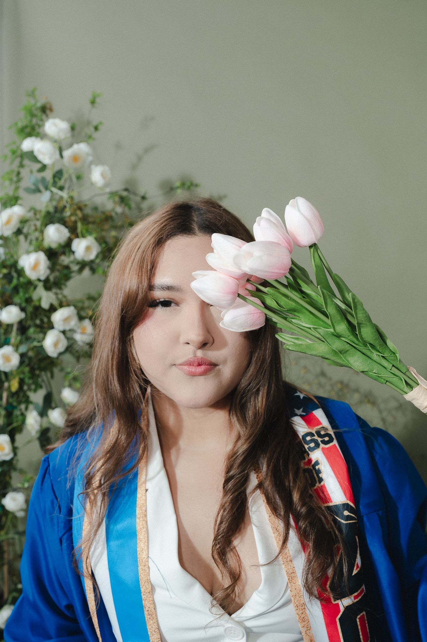 Young woman in blue graduation gown holds pink tulips near her face, with white flowers in the background.