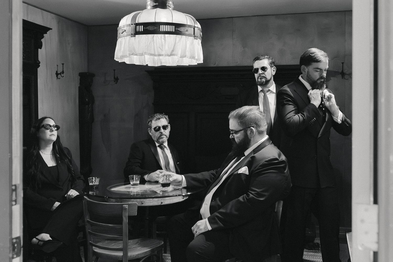 Black and white photo of five people in dark suits and sunglasses posed around a small table in a vintage room beneath a pendant lamp.