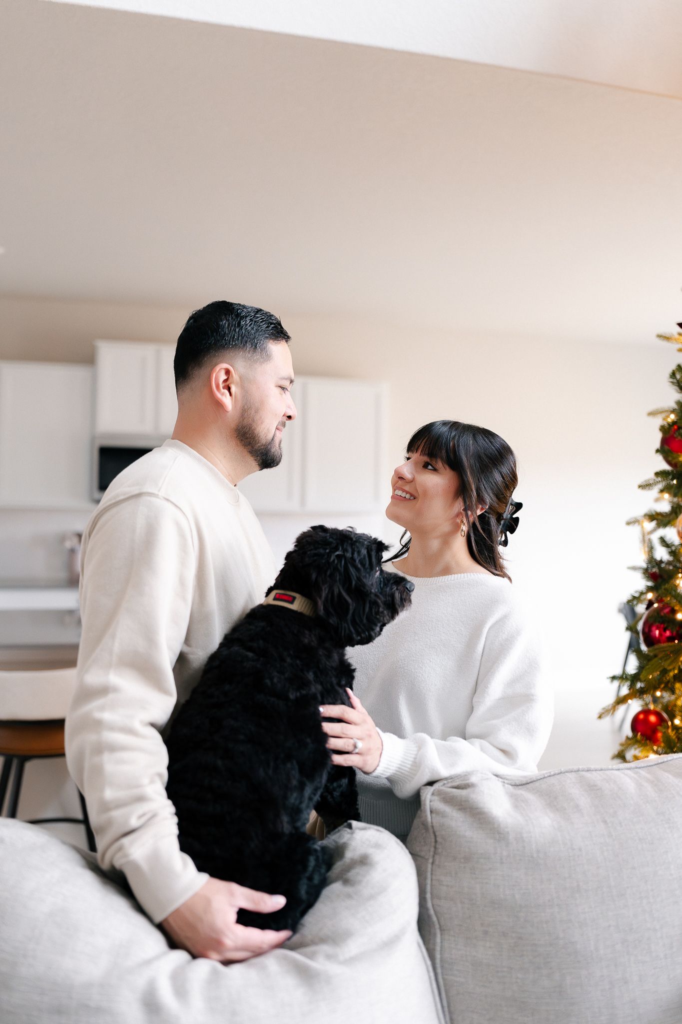 Couple in white sweaters sitting on couch with black dog during Christmas season with decorated tree in background.