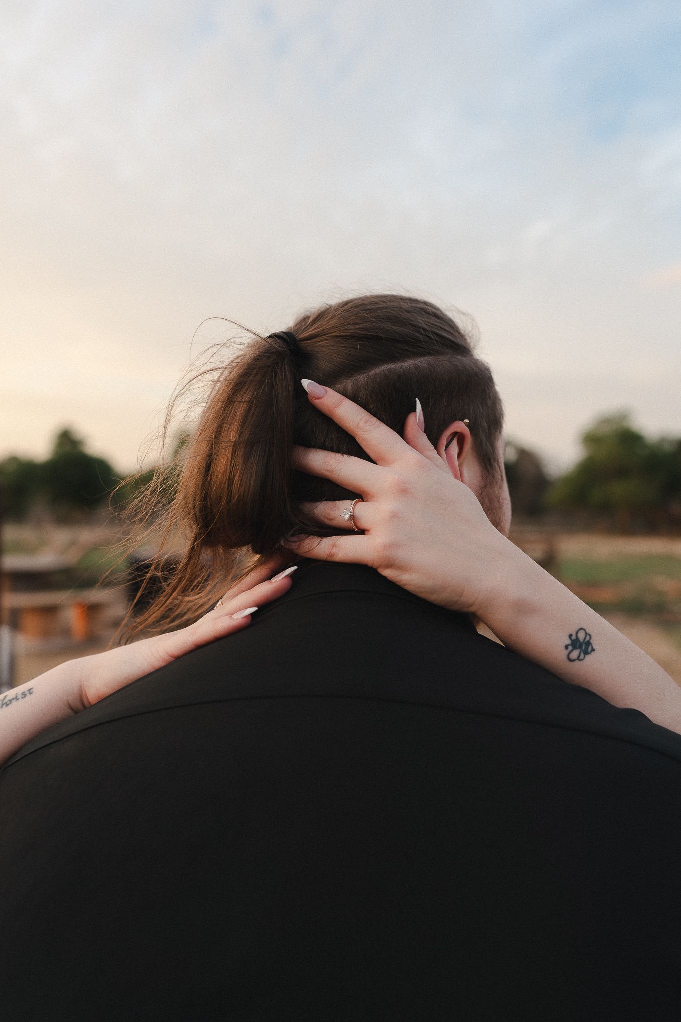 Close-up of a woman with tattoos embracing a man from behind in an outdoor setting at sunset.