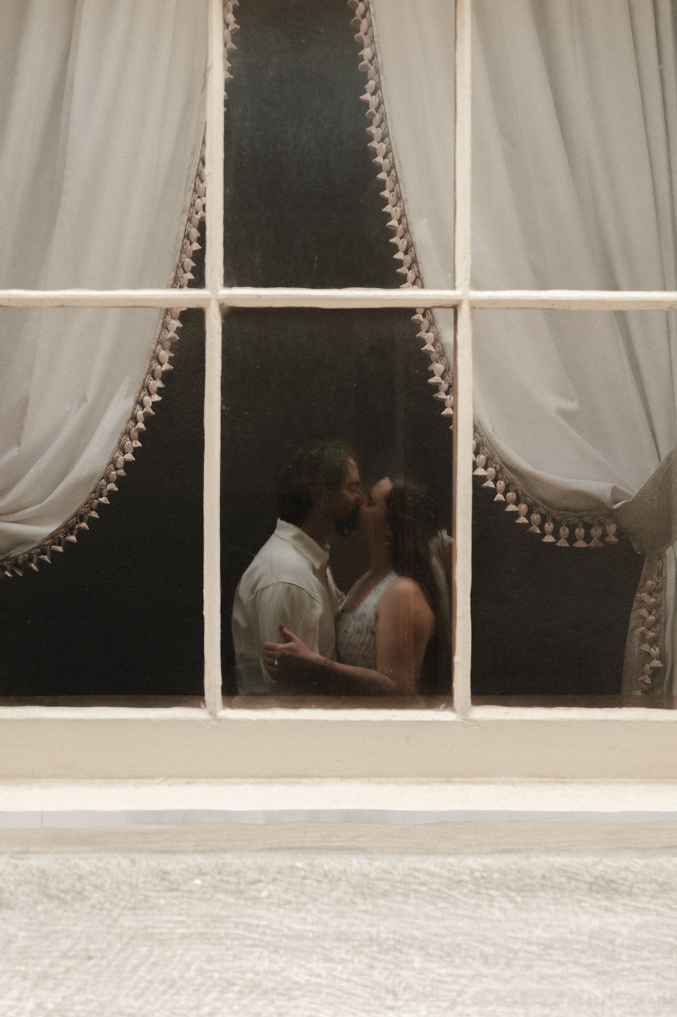 Couple sharing a romantic kiss visible through a large white-framed window with elegant curtains.