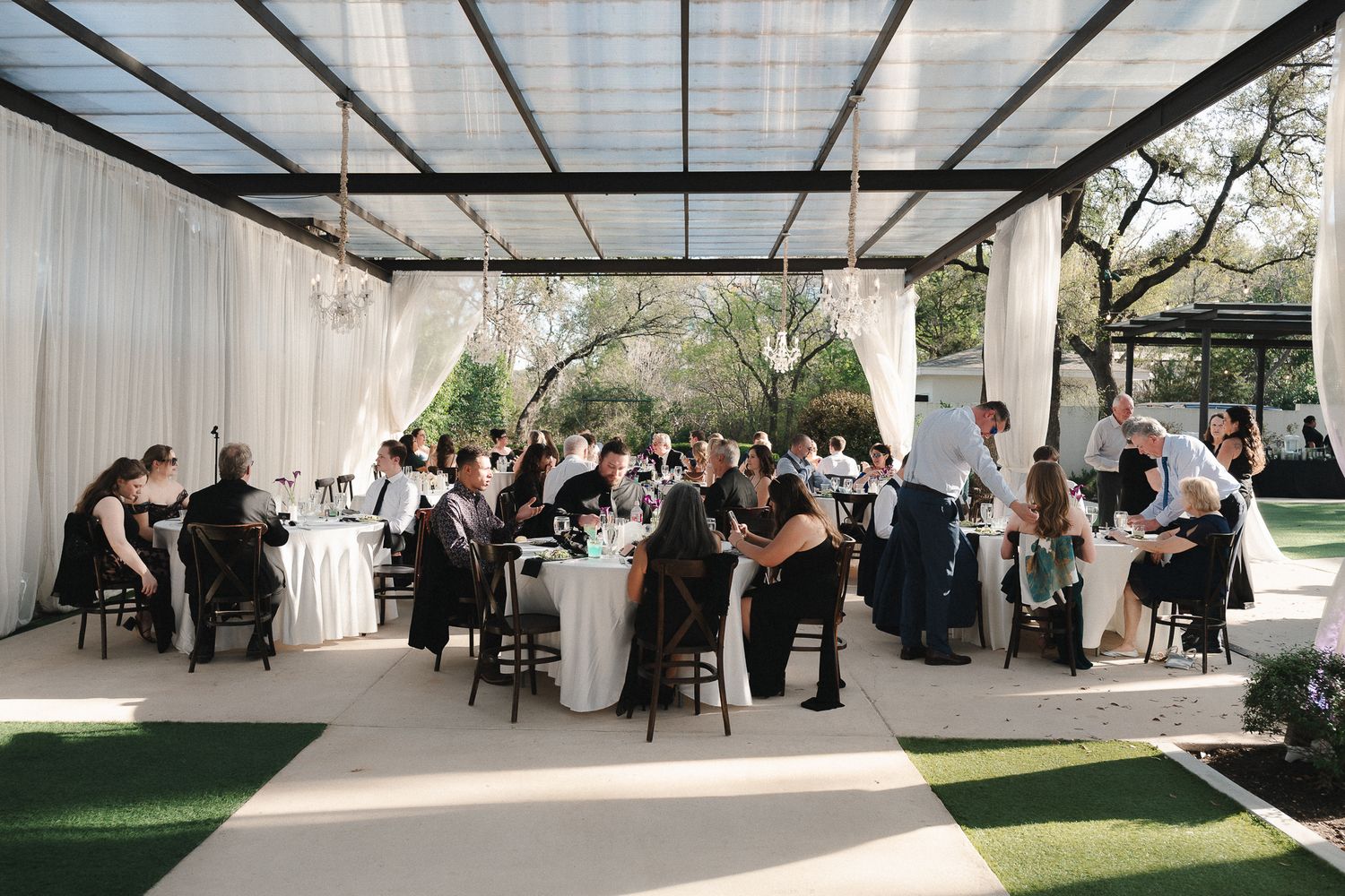 Elegant outdoor wedding reception under a pergola with white draping, chandelier lighting, and guests seated at round tables.