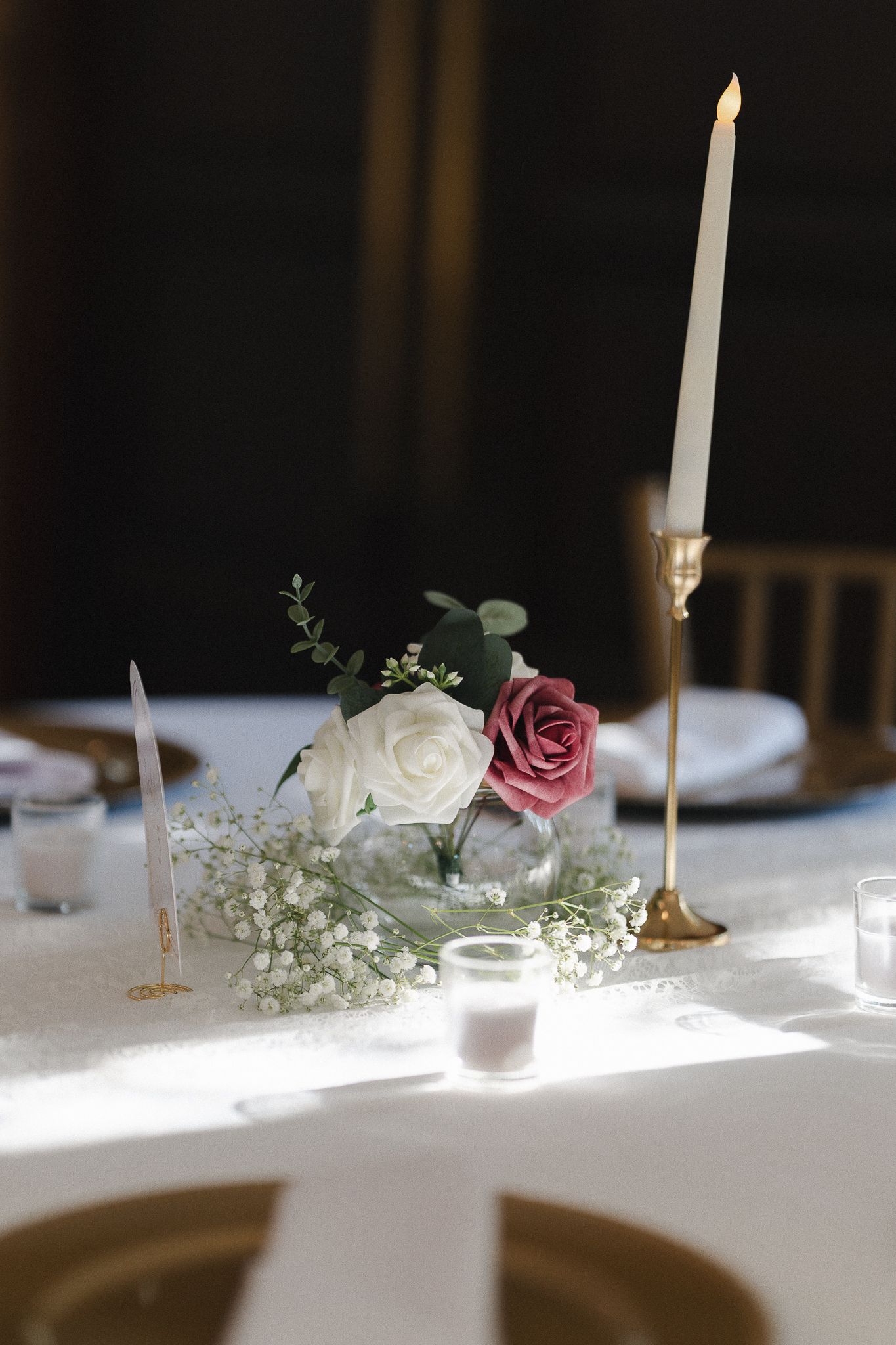 Elegant wedding table centerpiece with white and pink roses, baby's breath, gold candlestick, and lit taper candle on white linen.