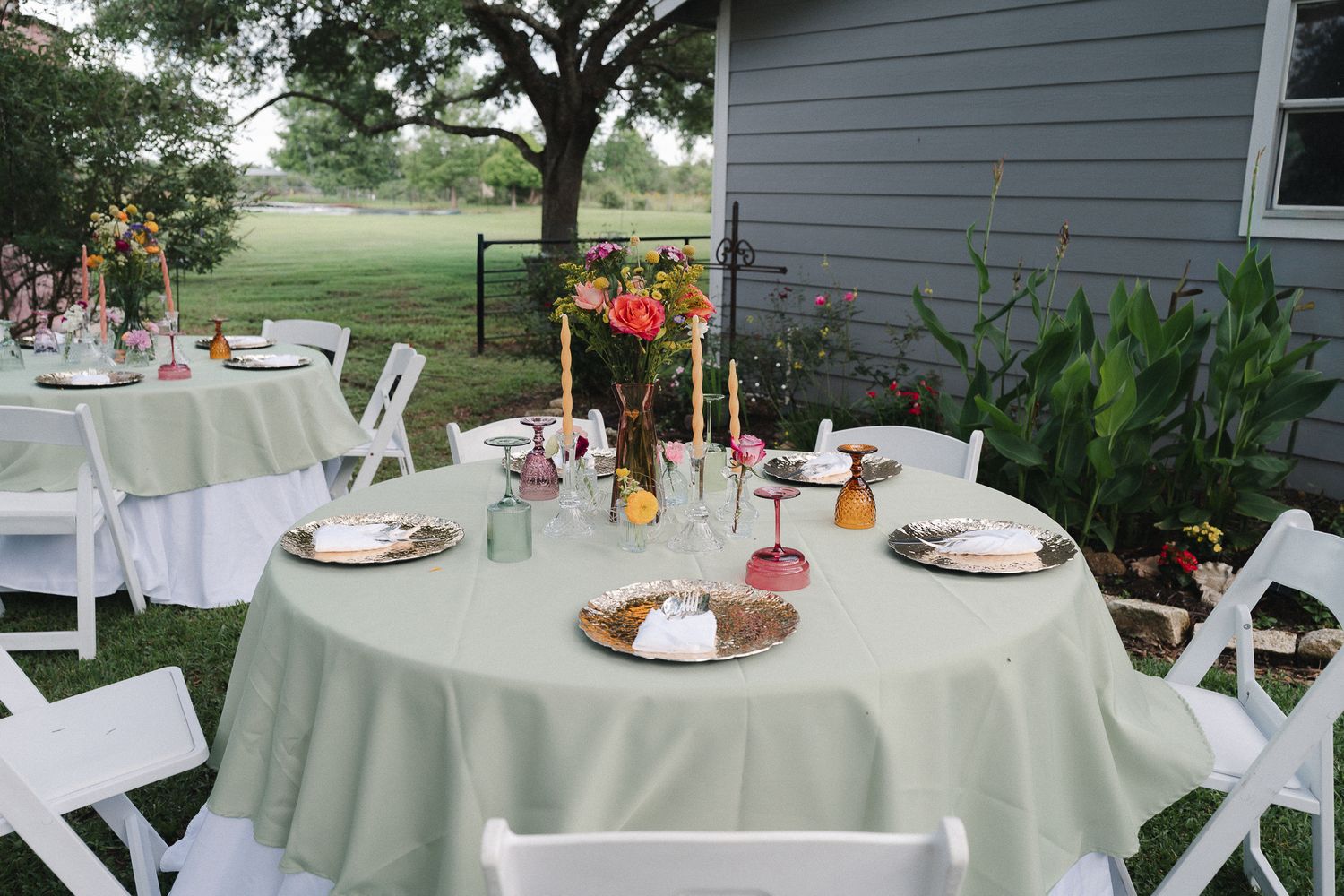 Outdoor wedding reception tables with sage green linens, colorful floral centerpieces, and white folding chairs on a green lawn.