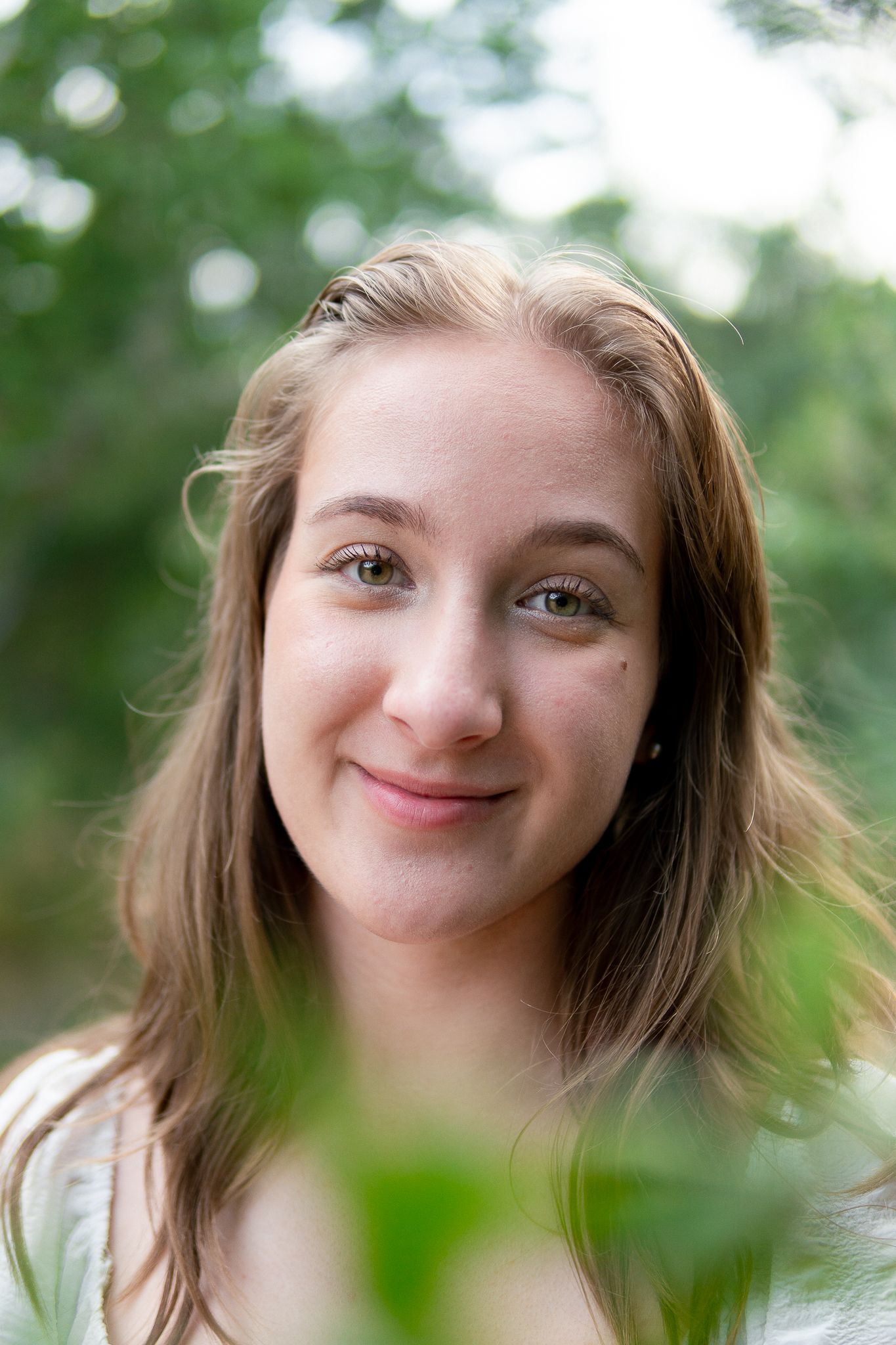 A person with brown hair smiles warmly at the camera through green foliage in a natural outdoor setting.