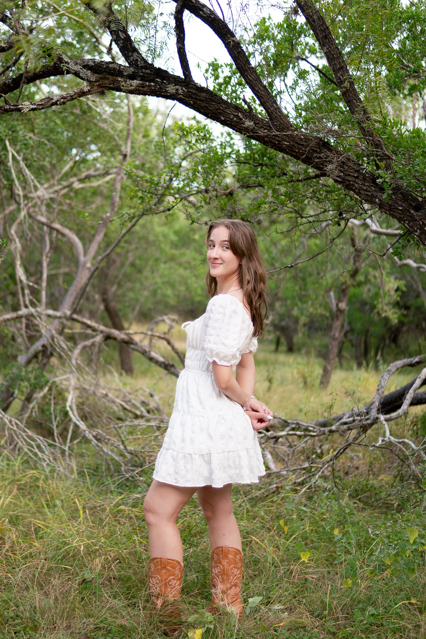 A person in a white lace dress and brown boots poses playfully in a natural wooded setting.