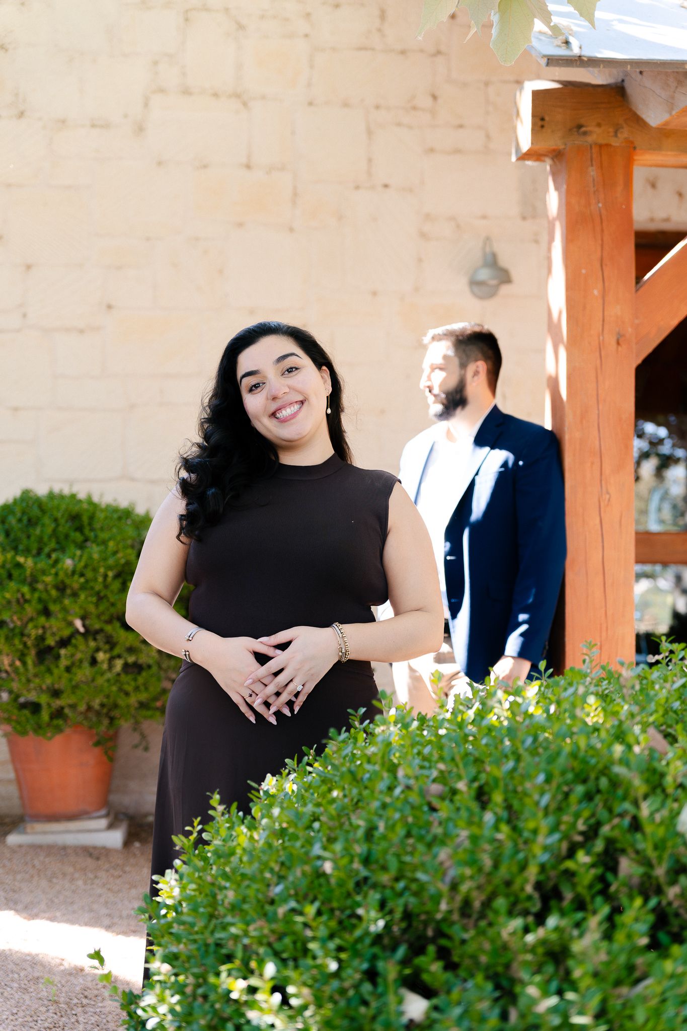 A couple poses outdoors near green shrubs and a beige wall with wooden post details.