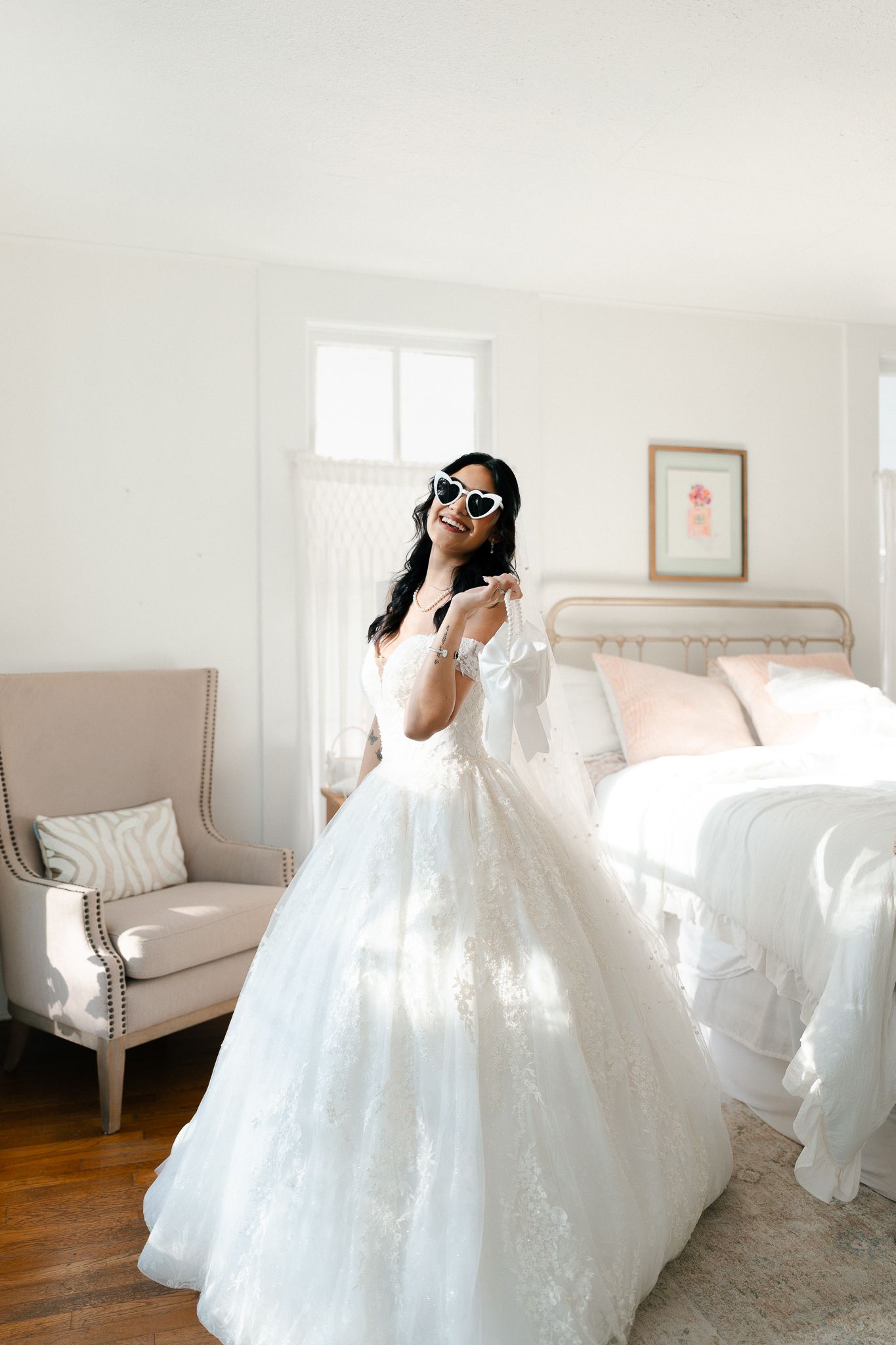 A bride in a voluminous white ballgown twirls in a bright sunlit room with neutral decor and hardwood floors.