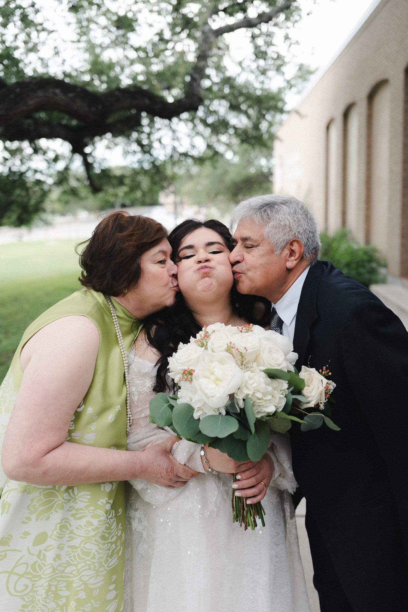 Bride holding white bouquet gets kissed on cheeks by parents in green dress and dark suit outdoors.
