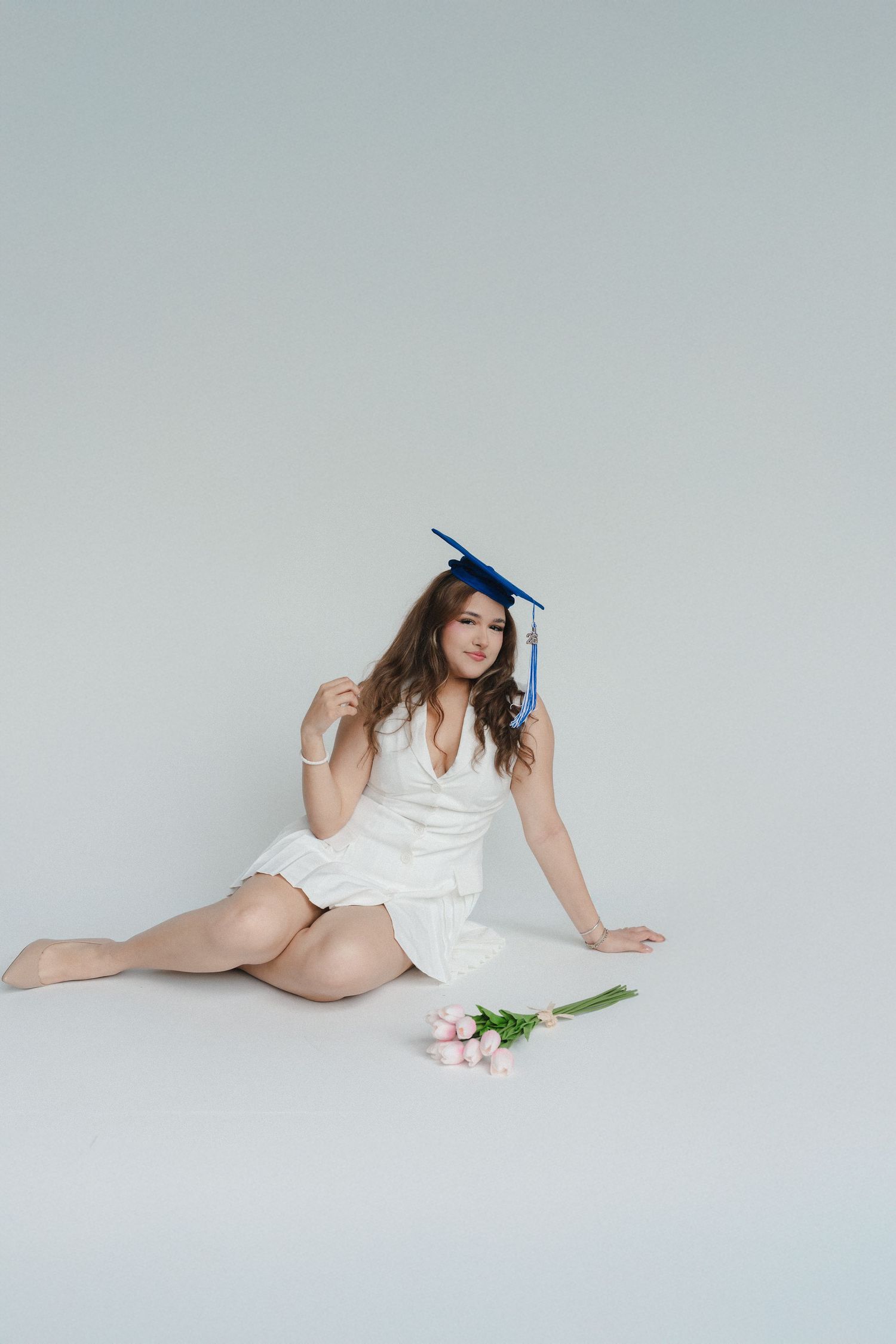 Young woman in white dress and blue graduation cap sits on floor with pink tulips in studio setting.