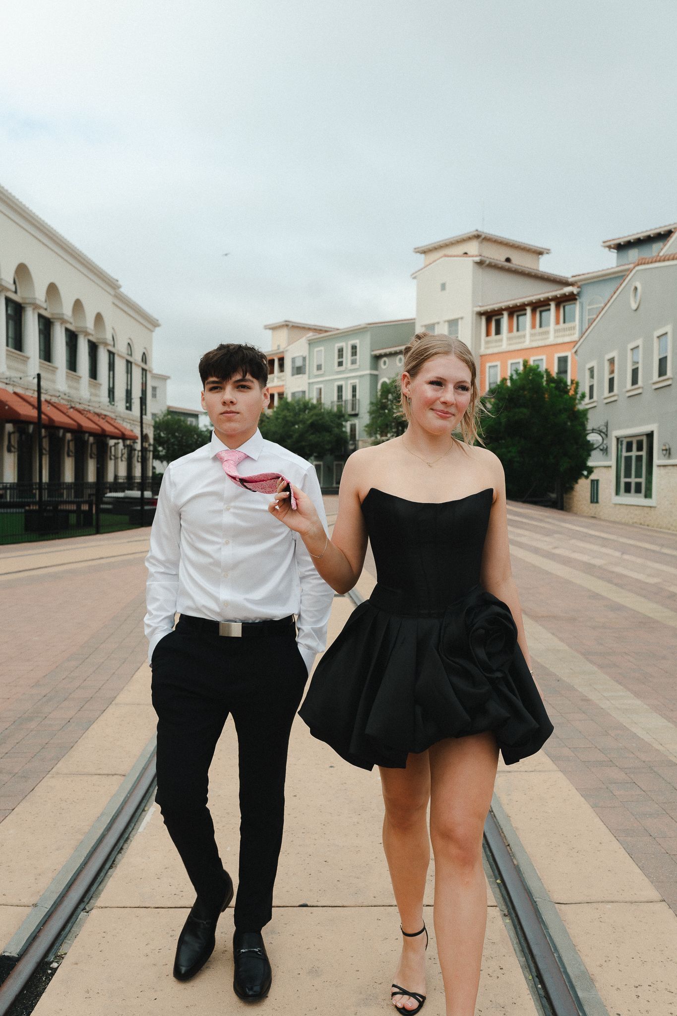 A young couple dressed in formal attire walks through an outdoor plaza, the girl in a black strapless dress.