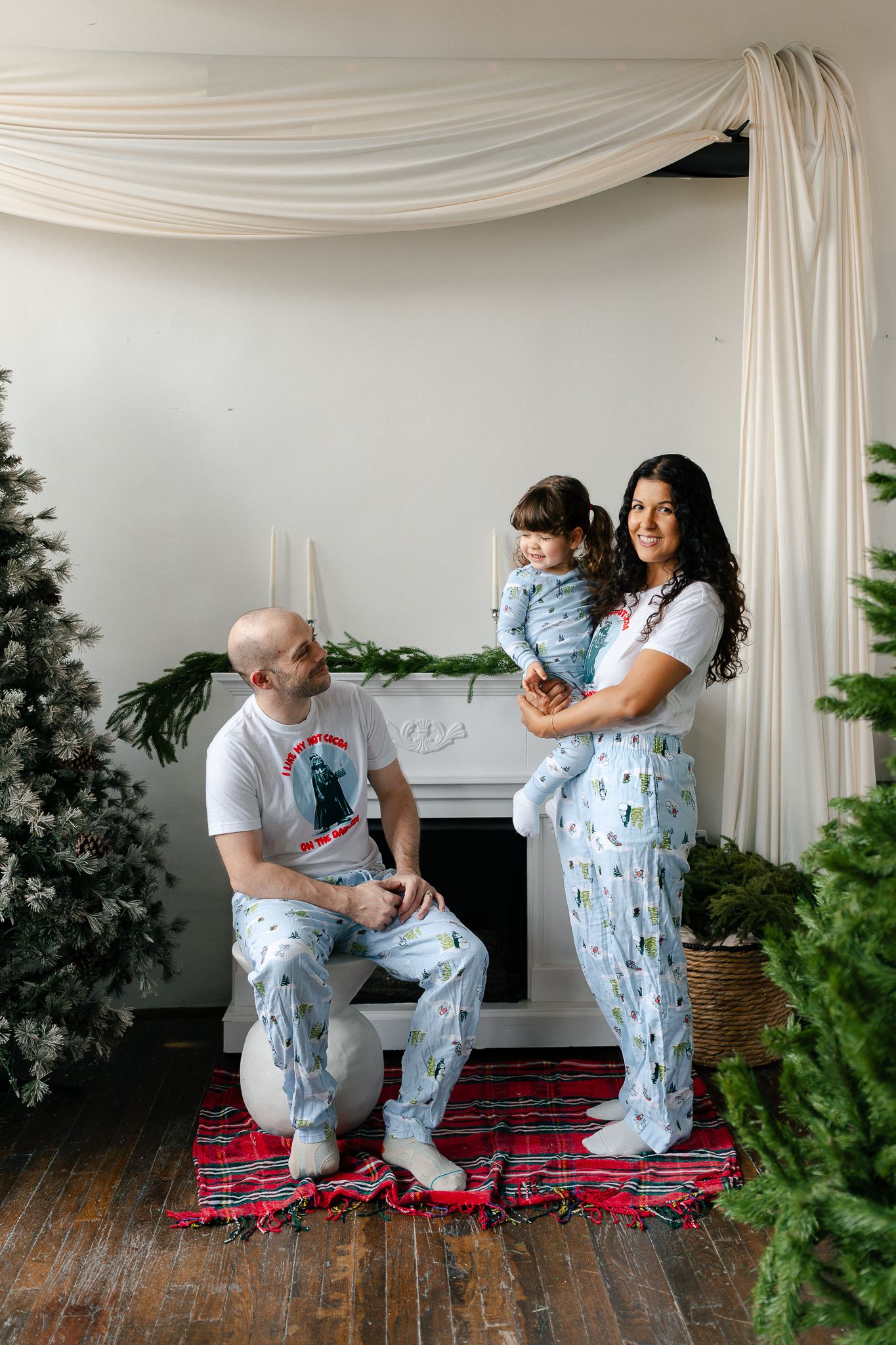A couple wearing matching blue floral pajamas share tender moments in a studio setting with white drapes and a red plaid blanket.