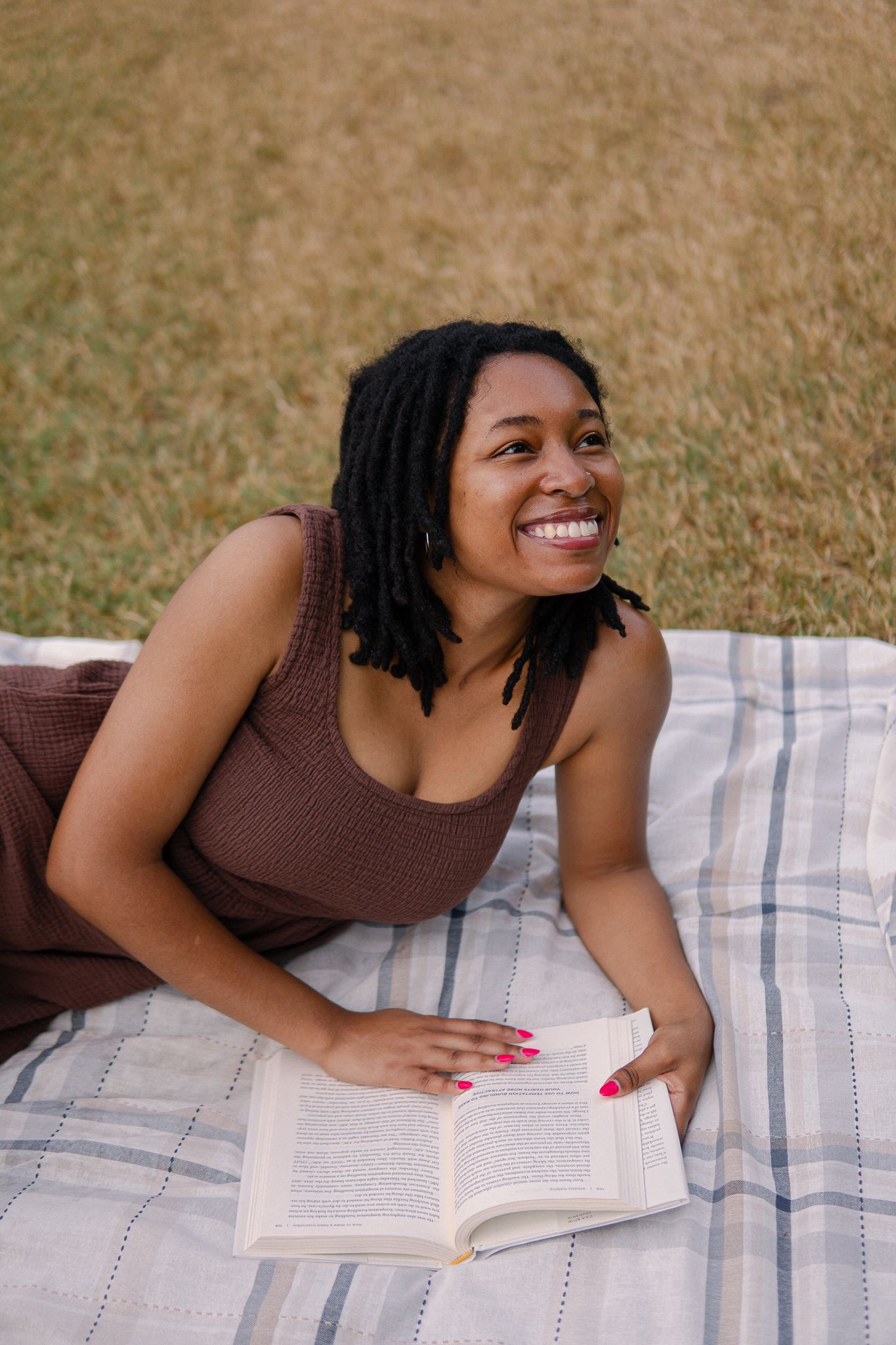 Person reading a book while relaxing on a plaid blanket outdoors on a sunny day.