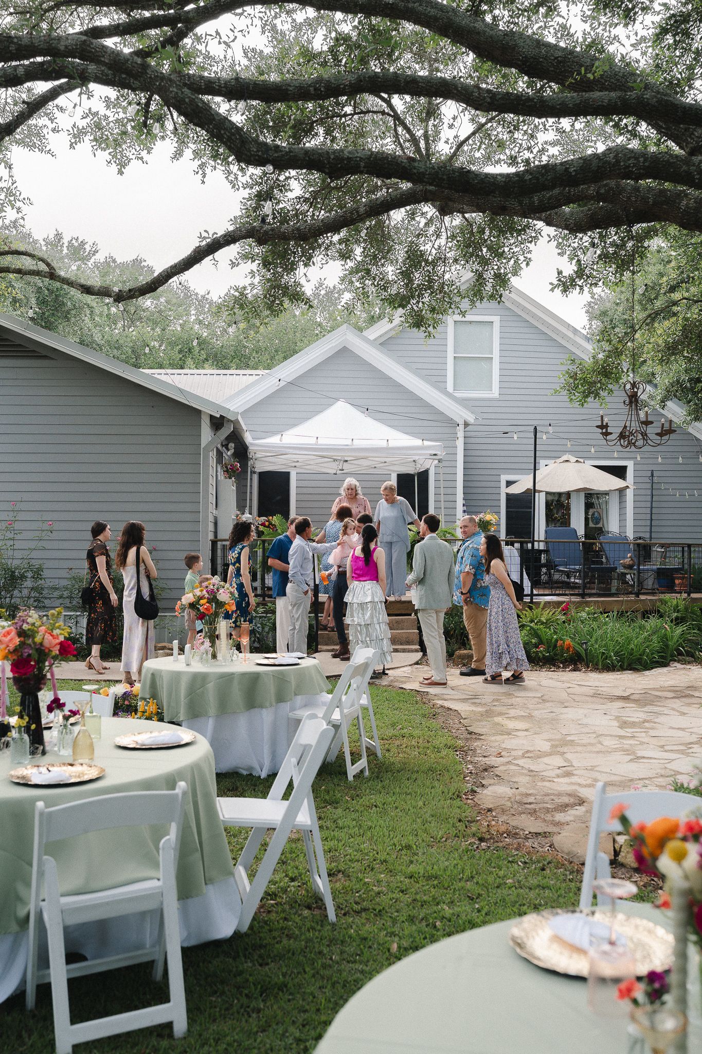 Outdoor garden party with guests mingling on a patio, white round tables, and a gray farmhouse venue surrounded by trees.