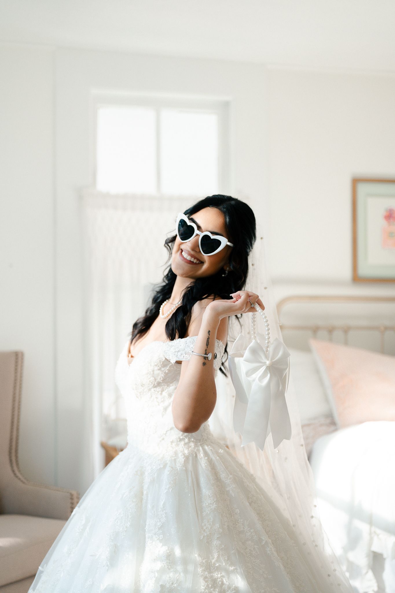Smiling bride in white lace ballgown and heart-shaped sunglasses holding pearl accessories in bright bedroom.