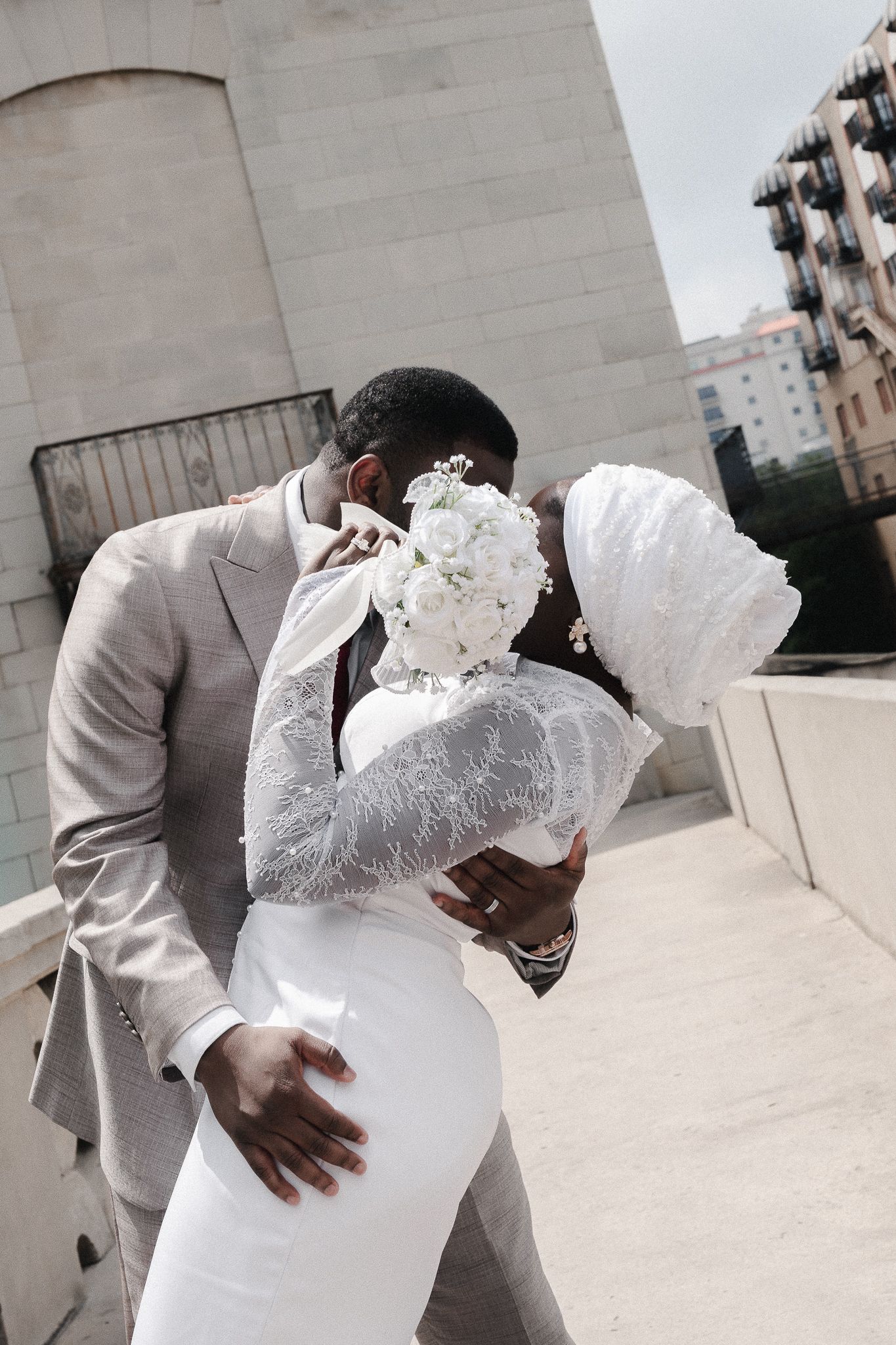 A Black couple shares a romantic kiss on their wedding day, bride in white lace gown with bouquet, groom in beige suit.