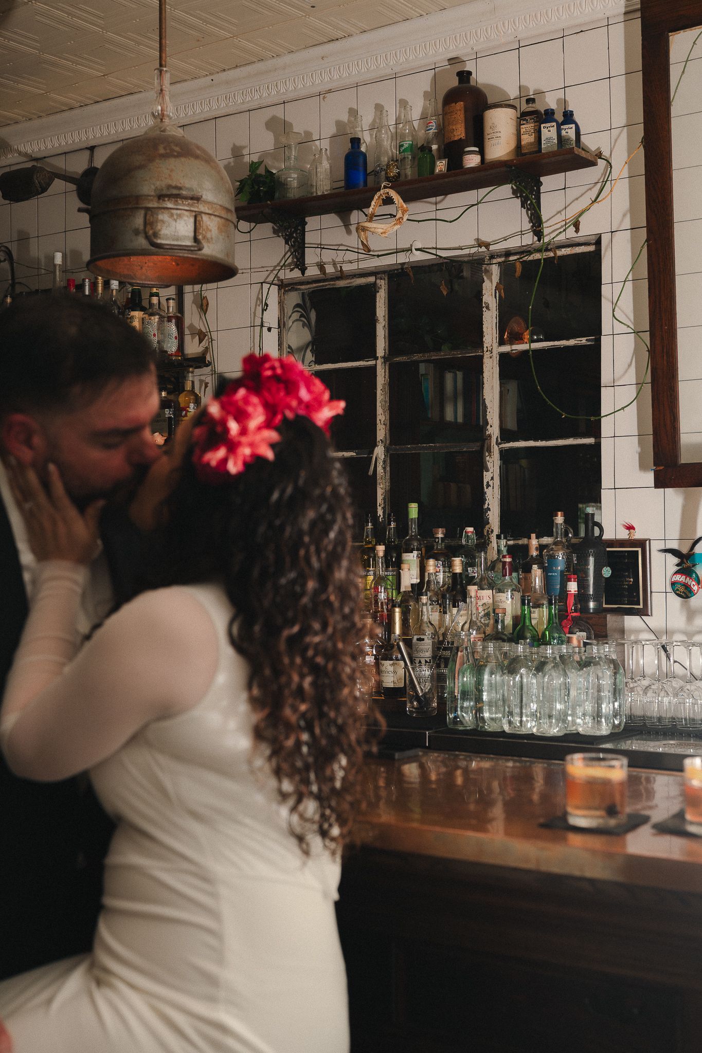 Bride and groom share a romantic kiss at a bar, bride wearing white dress with pink flowers in curly hair.
