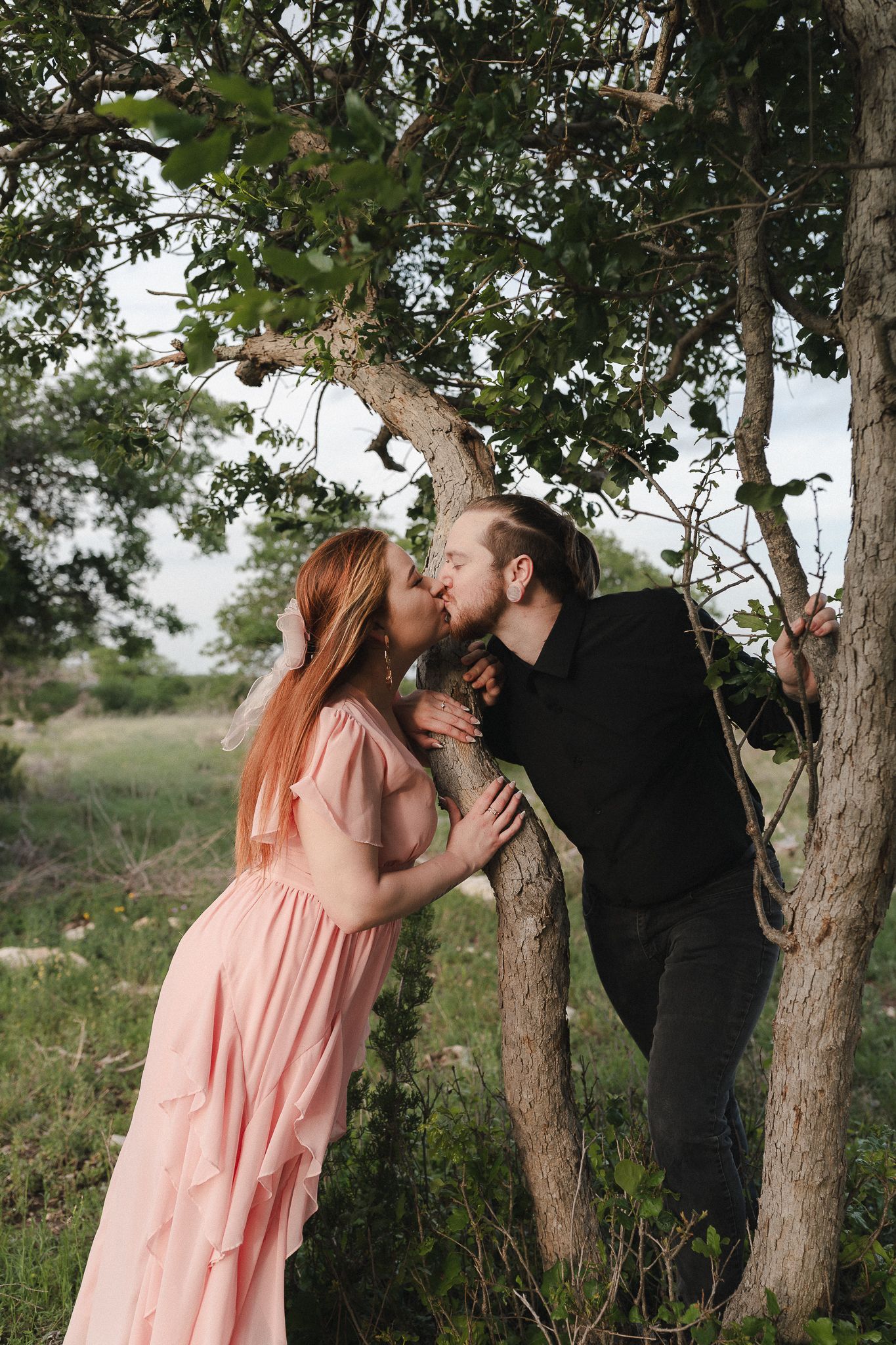 Couple kissing romantically beside a tree in a lush green field, woman in pink dress, man in black outfit.