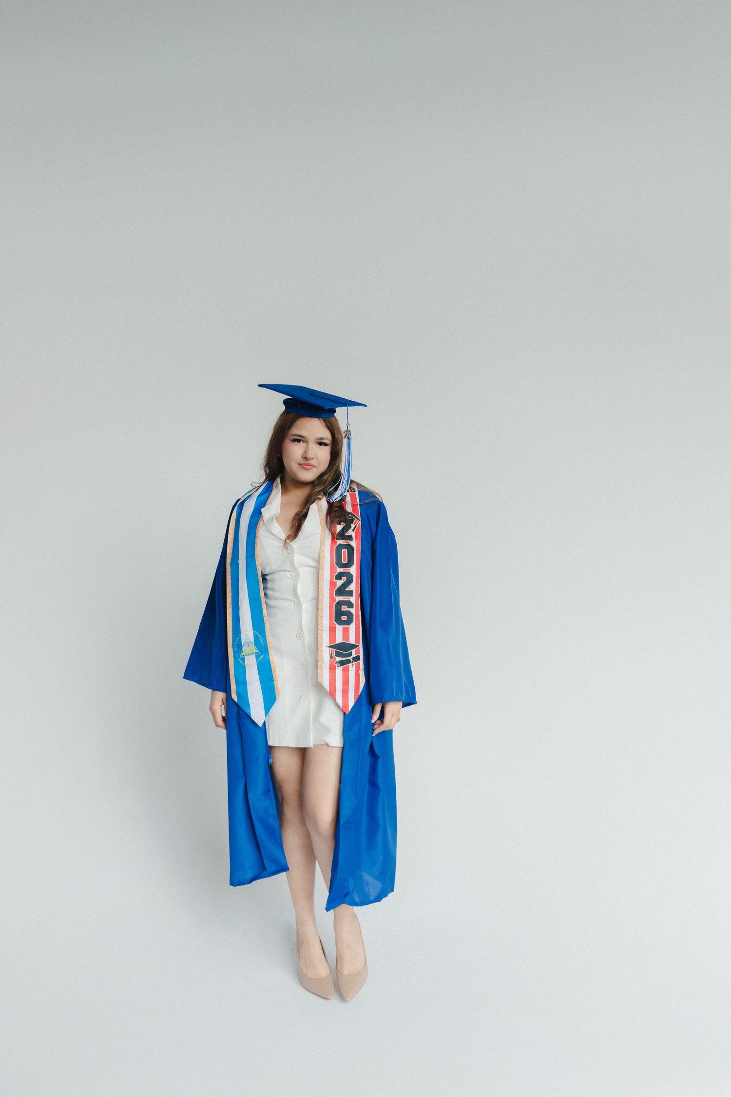 Young woman in blue graduation cap and gown with honors sash, posing confidently against white background.