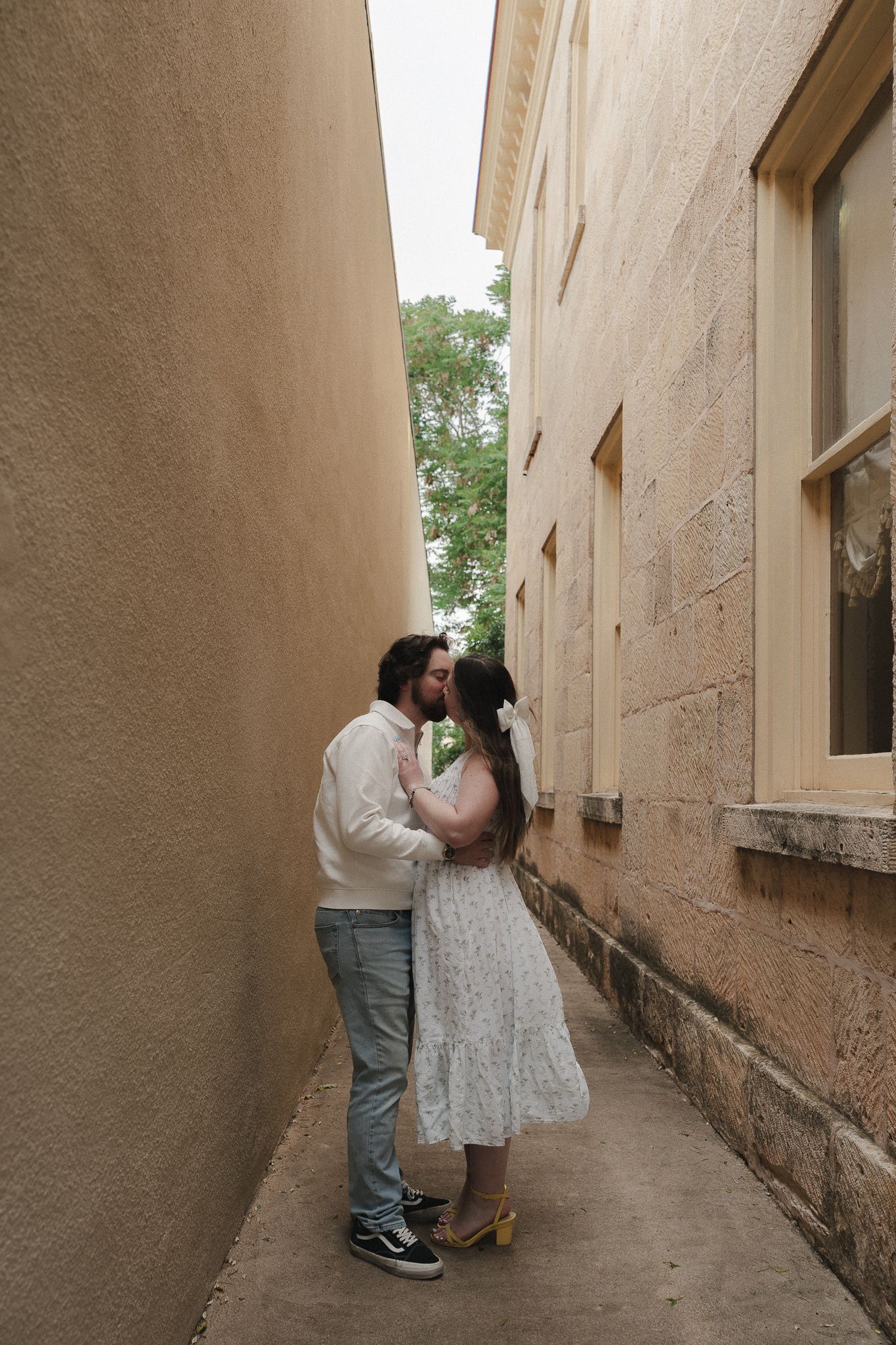Couple sharing a romantic kiss in a narrow alleyway between two stone buildings, woman in white dress.