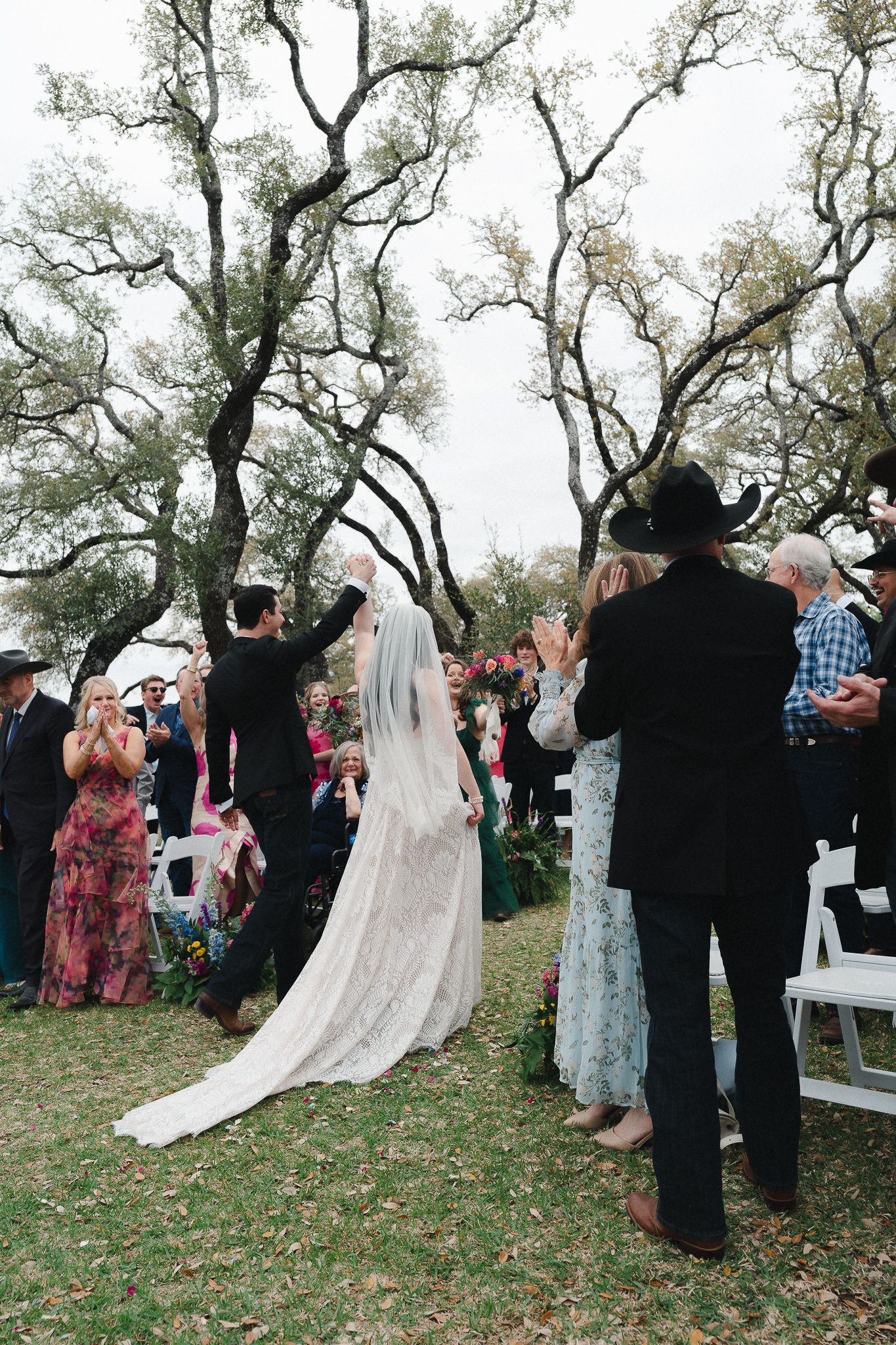 Bride and groom raise hands at outdoor wedding ceremony under oak trees with guests applauding.