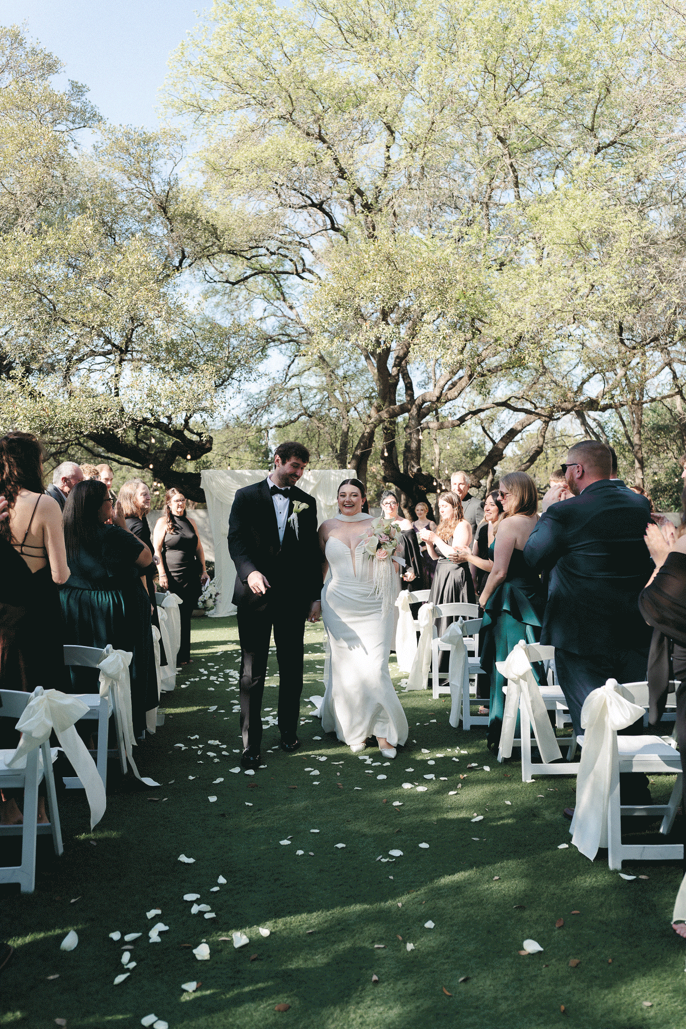 Bride and groom walk down outdoor aisle after wedding ceremony, guests cheering, white chairs, rose petals, lush trees.