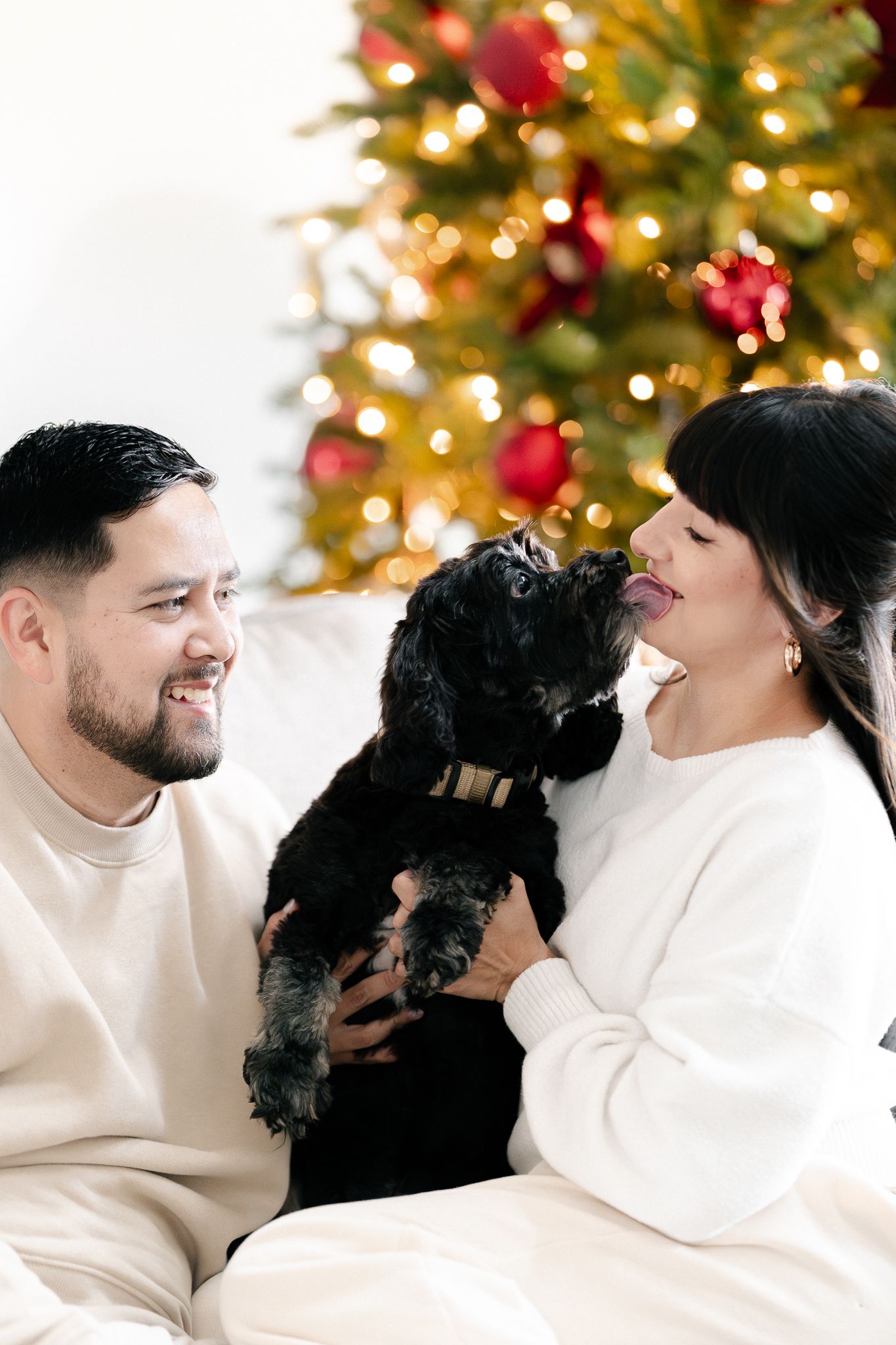 A couple in white sweaters share a romantic moment in front of a Christmas tree with twinkling lights and red ornaments.