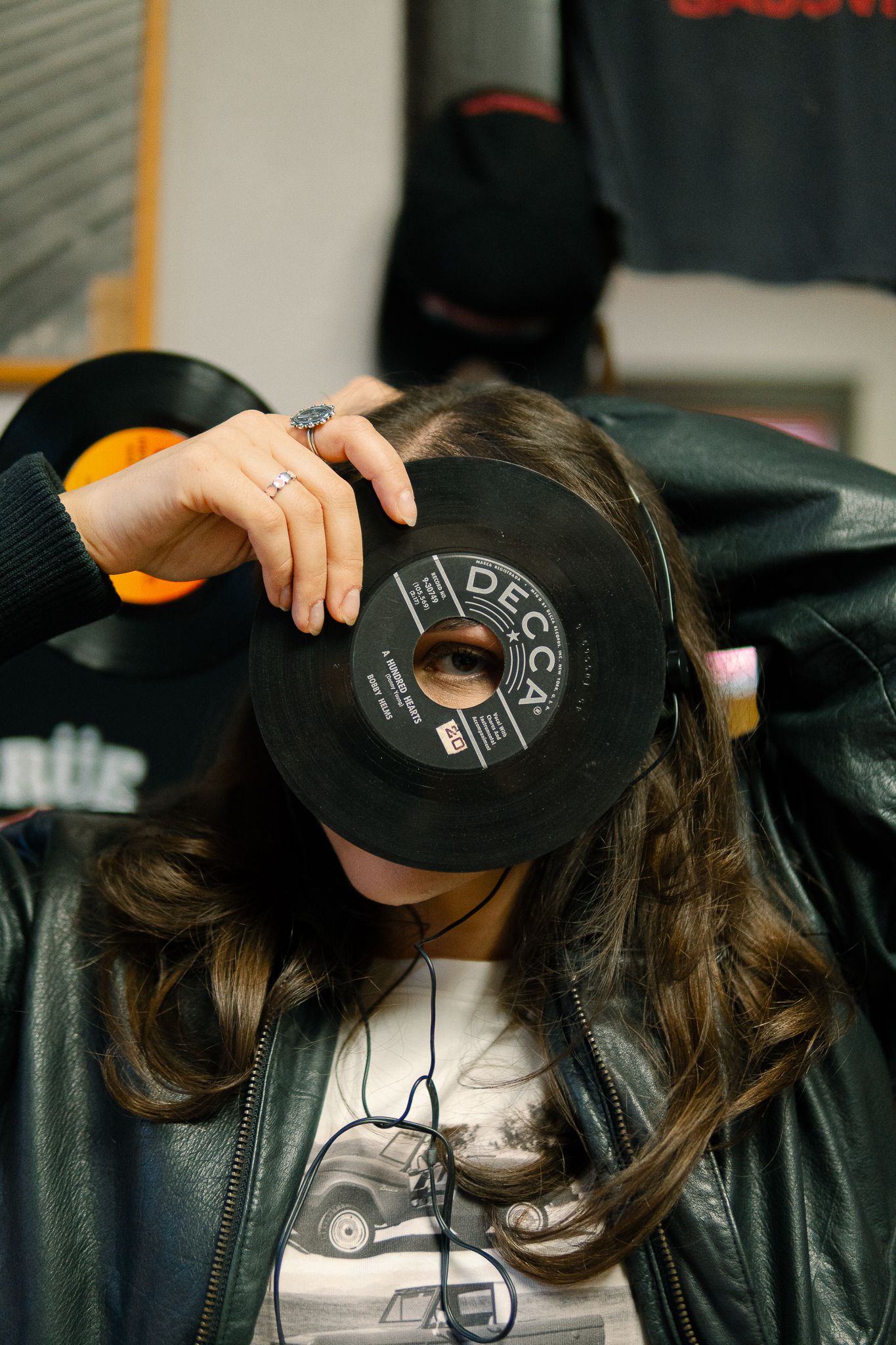 Person playfully holds up a vinyl record in front of their face while wearing a leather jacket and graphic t-shirt.