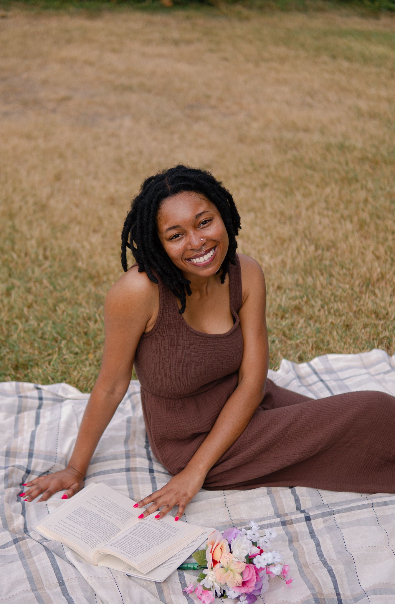 A person in a brown outfit lies on a picnic blanket with flowers while reading outdoors on a sunny day.