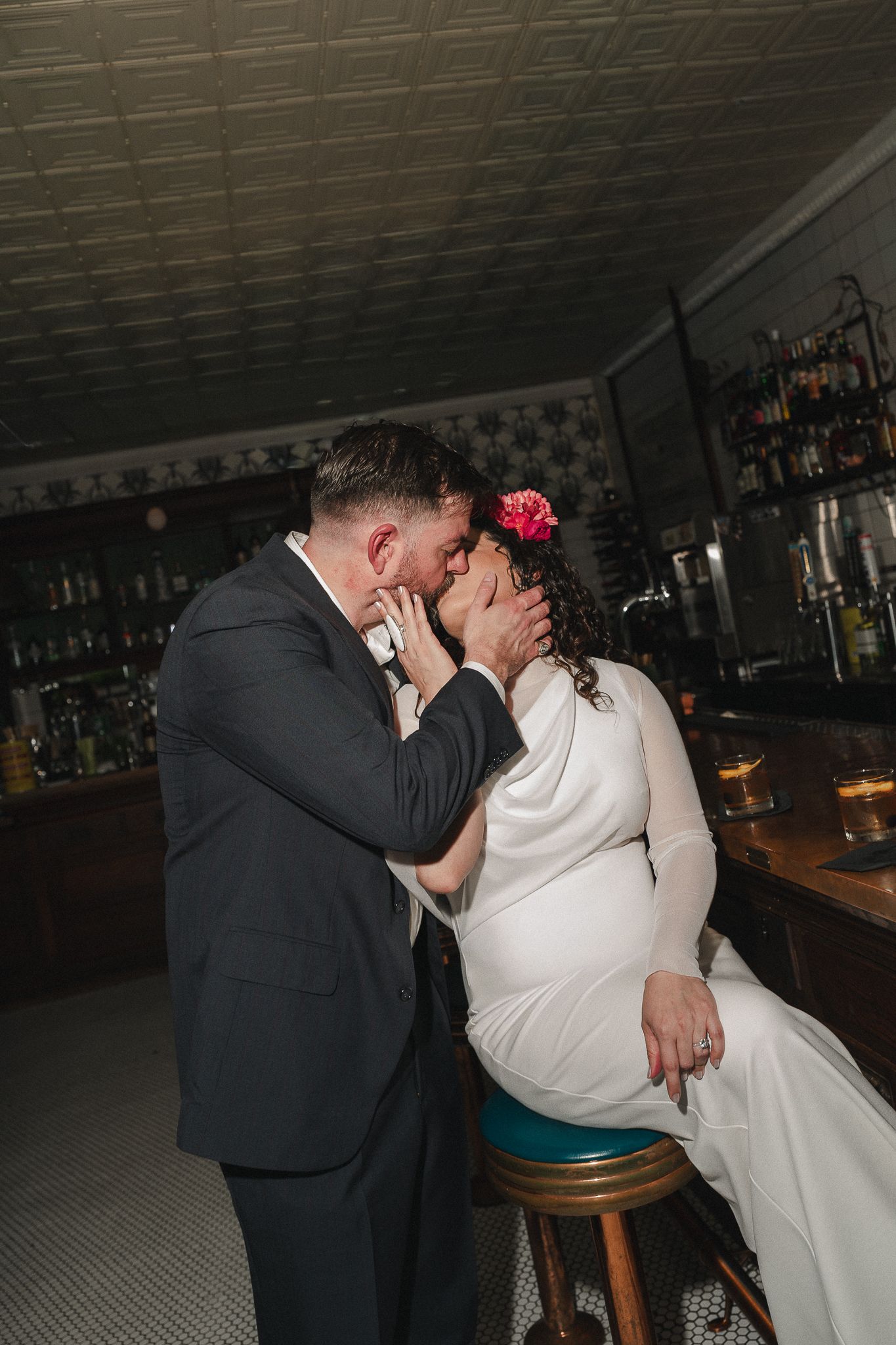 A groom in a dark suit tenderly kisses his bride in a white gown at a bar, as she sits on a stool with a pink flower in her hair.