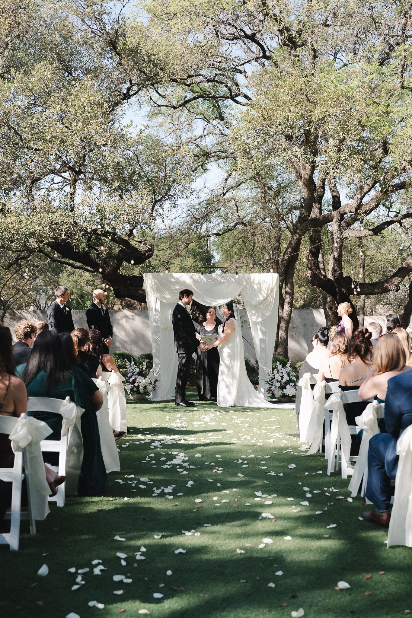 Outdoor wedding ceremony under oak trees with white aisle, floral arch, and guests seated in white chairs on green lawn.