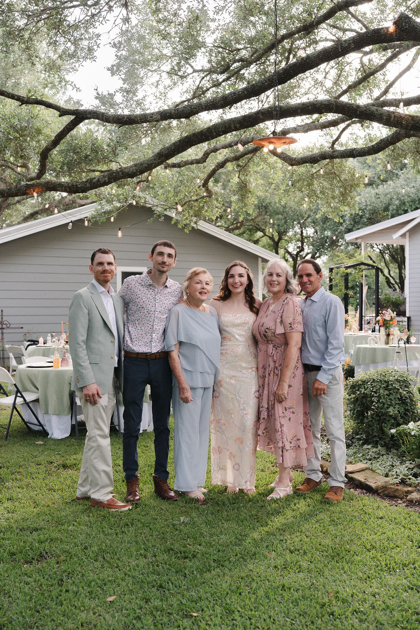 Family group photo at an outdoor wedding reception, posing under oak trees near decorated tables.