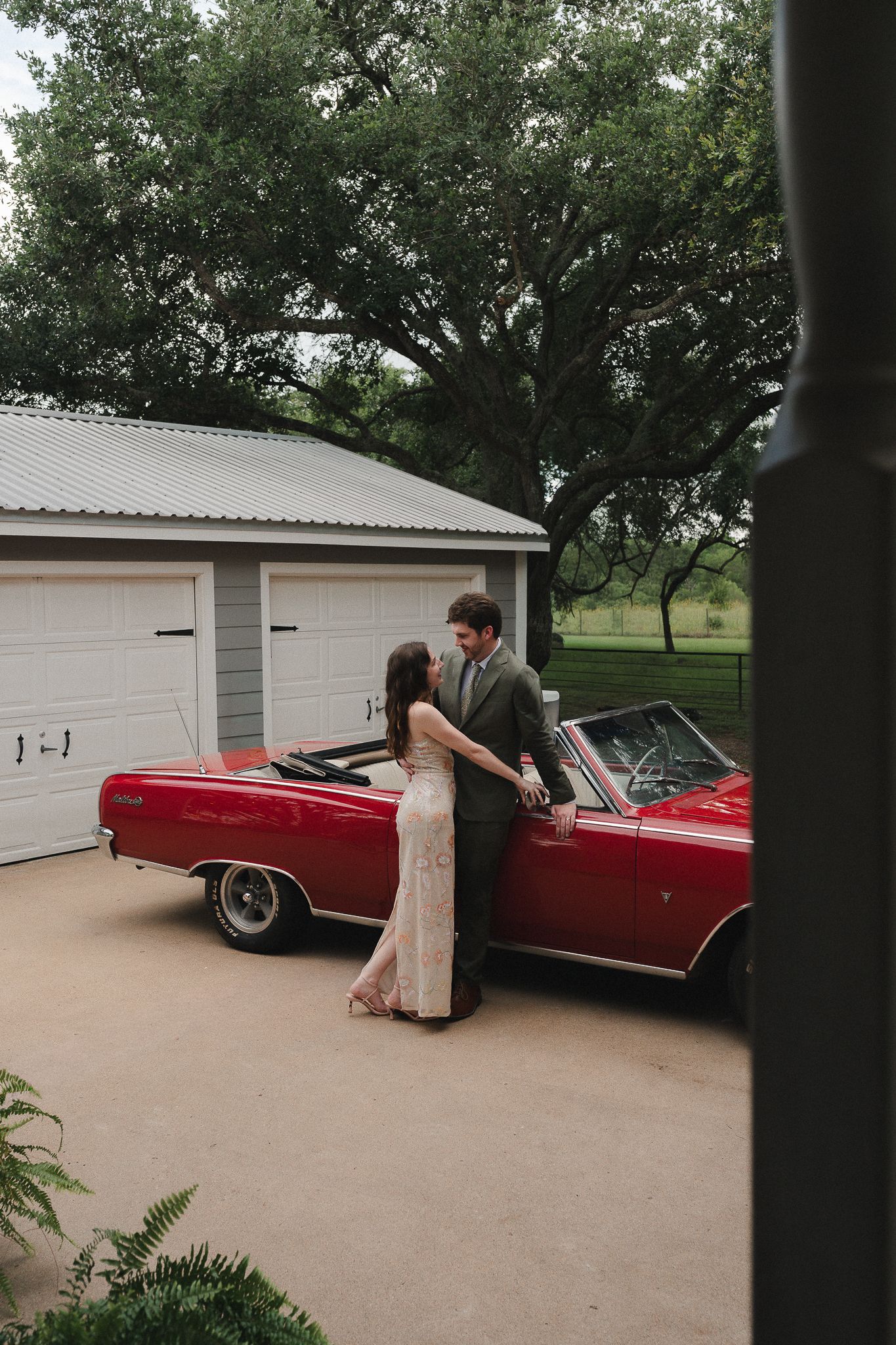 Couple sharing a romantic moment beside a classic red convertible in a driveway with a white garage and lush trees.