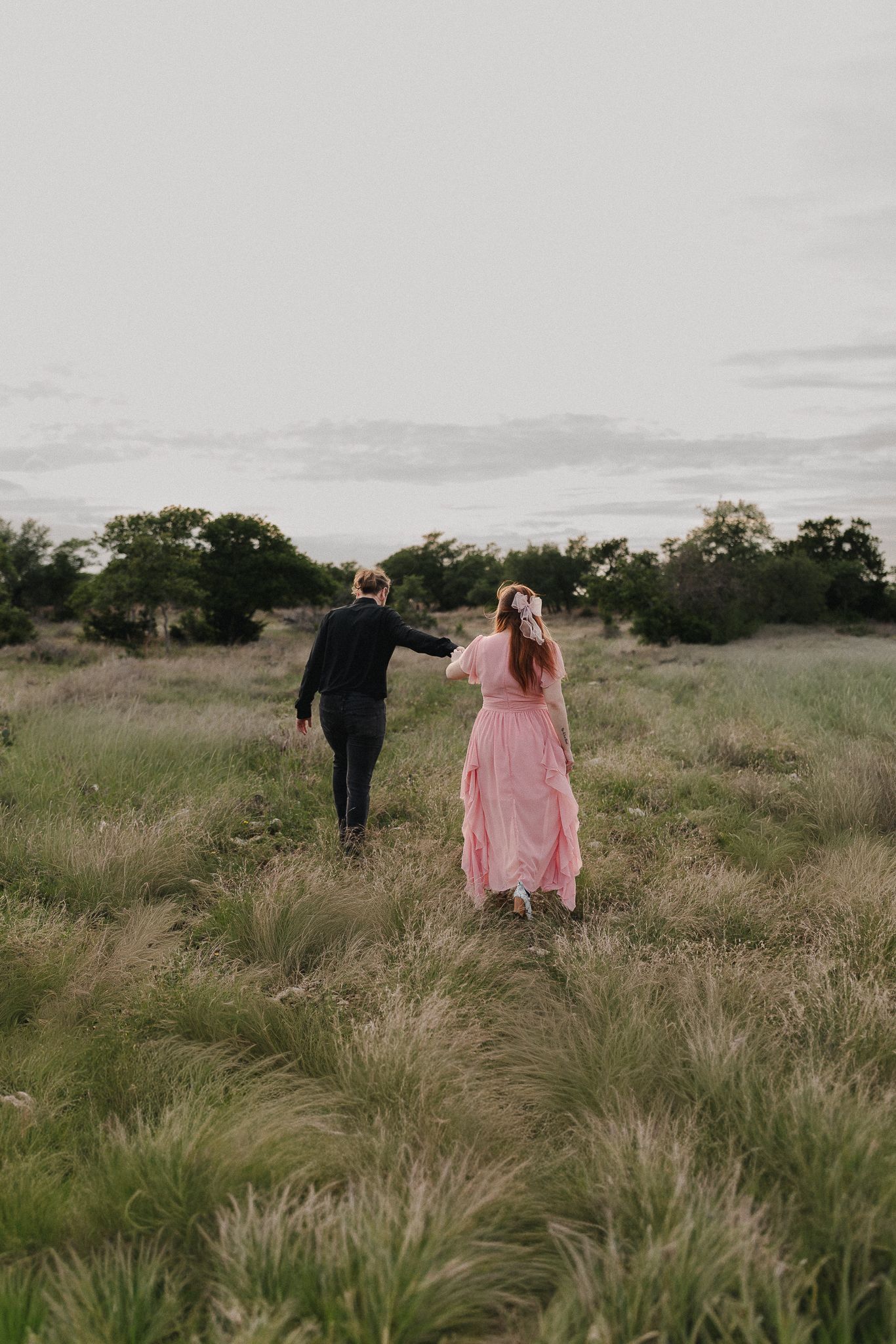 A couple walks hand-in-hand through a lush green field, woman in pink dress, man in dark suit, trees in background.